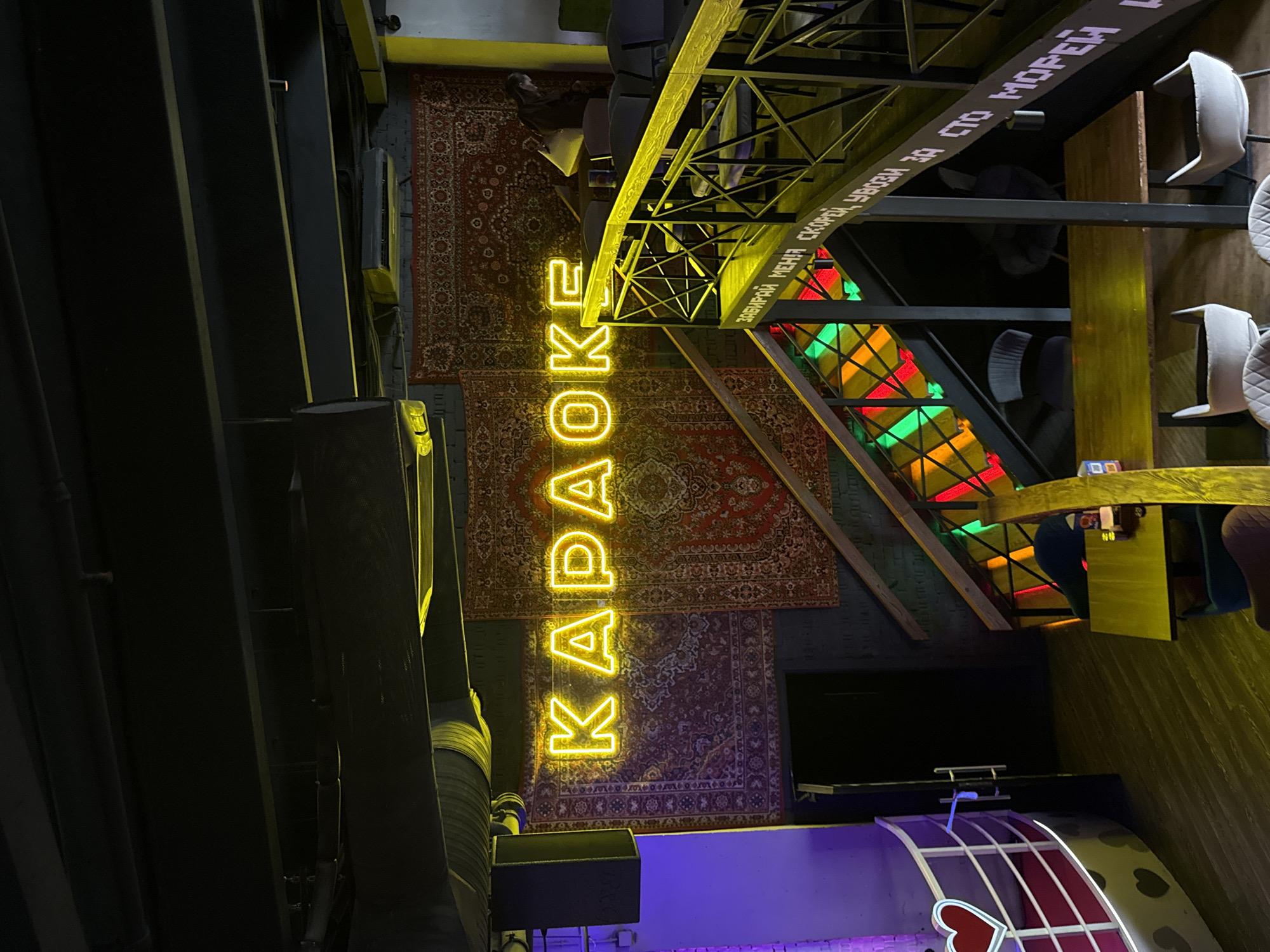 The image shows the interior of a karaoke bar in Moscow, Russia. A vertical neon sign spells out "KARAOKE" in yellow. The floor is covered with patterned rugs, and the bar features a steel structure with a sign in Russian that reads, "ЗАБИРАЙ МЕНЯ СКОРЕЙ УВЕЗИ ЗА СТО МОРЕЙ" ("Take me away quickly beyond a hundred seas"). A stained glass window with red, green, and yellow panes is visible. The bar has wooden tables and stools. There is also a stage with neon lighting in the lower portion of the image.