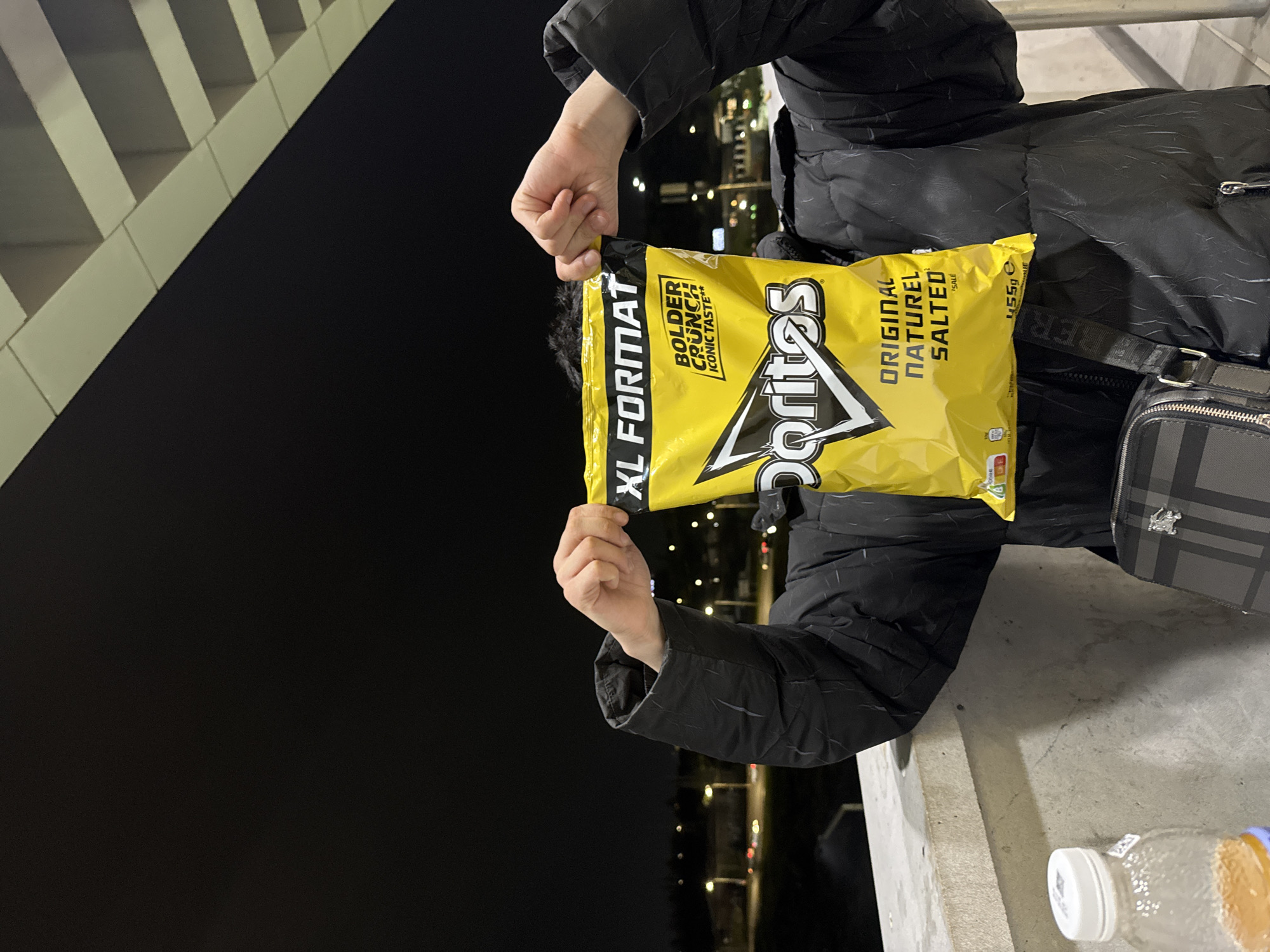 A person wearing a black puffer jacket holds a bag of Doritos chips in front of their face at night. The chips are "XL Format" and "Original Naturel Salted". The person stands next to a concrete ledge, and a water bottle sits on the ledge to the right of the person. Lights are visible in the distance. The photograph appears to have been taken at night. It is not possible to discern the specific location within the city of Yerevan, Armenia, based on the provided image.