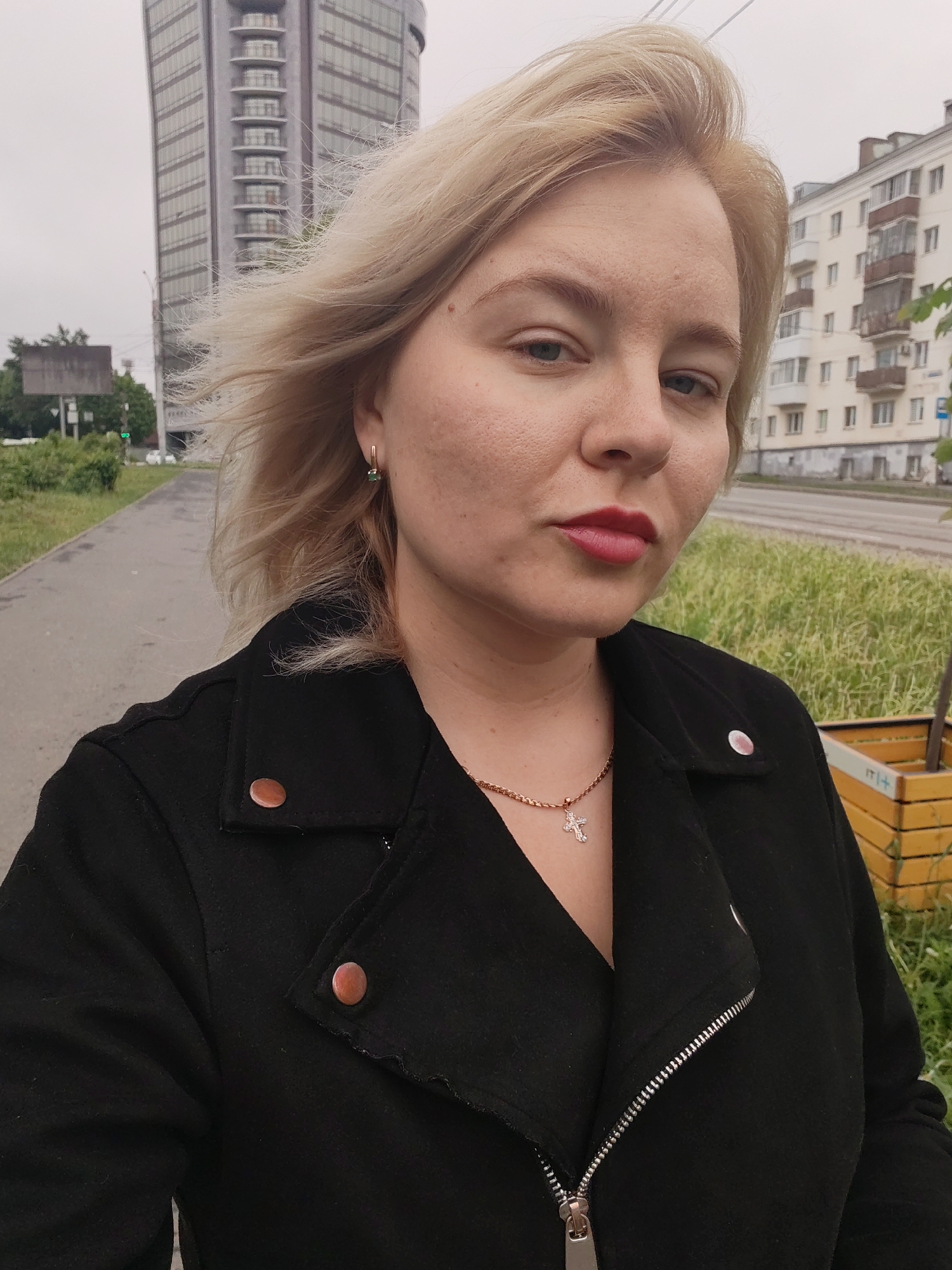 A woman with blonde hair, wearing a black jacket and a cross necklace, is standing outside on a paved path in Izhevsk, Russia. A tall building is visible in the background. The path has grass on both sides. The sky is overcast.