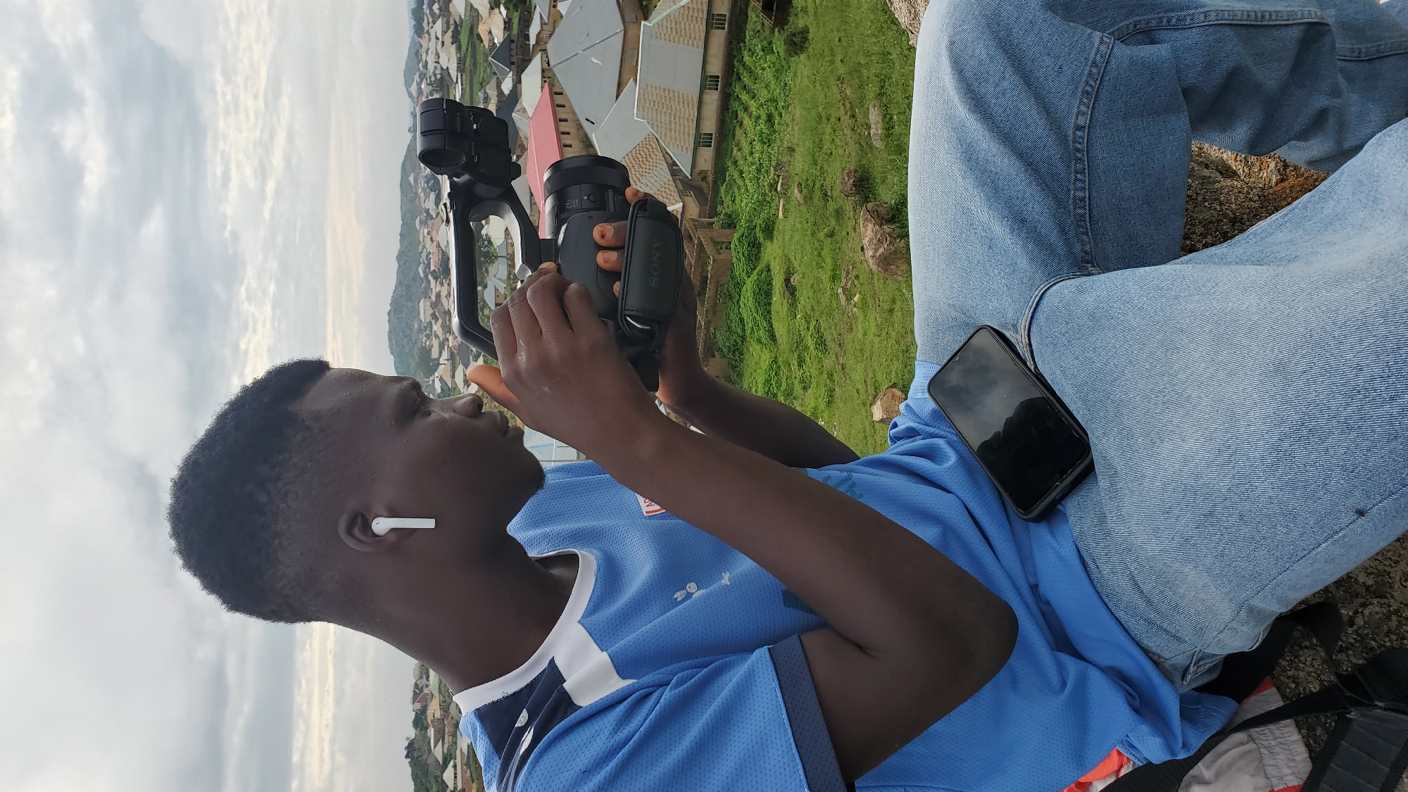 A young Black male with short, dark hair and white earbuds is seated on a rock outcrop, holding a Sony camcorder with both hands and looking through the viewfinder. He is wearing a bright blue short-sleeved t-shirt with white and dark blue trim on the collar and sleeves, and light blue denim jeans. A black smartphone with a black case is resting on his lap, screen up. In the background, a dense residential area with a mix of modern and older homes with various colored roofs stretches out towards a hazy horizon. Green grass and scattered rocks are visible on the outcrop. The sky is overcast with a grey and white cloudy texture. This scene appears to be on an elevated position overlooking a section of Jos, Nigeria.
