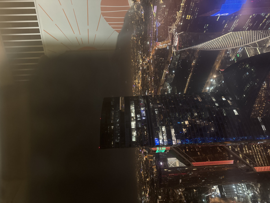 The scene depicts a high-angle nighttime view of an illuminated urban landscape, located in the city of Khimki, Russia. Prominently featured are several modern skyscrapers, brightly lit against a dark sky. The central structure is a tall, dark-glassed building with numerous illuminated windows, crowned by faint red beacon lights. To its lower right, a distinct helical or spiraled skyscraper is visible, its white facade catching the city lights. Another multi-story building to the left displays vertical red and green lighting strips. Below, a river flows, bordered by illuminated roads and infrastructure, with city lights extending extensively into the hazy distance. The foreground right edge shows a partial view of an interior space, including a white vertical railing and a beige wall adorned with a red-orange sunburst mural. No individuals or specific actions are discernible.