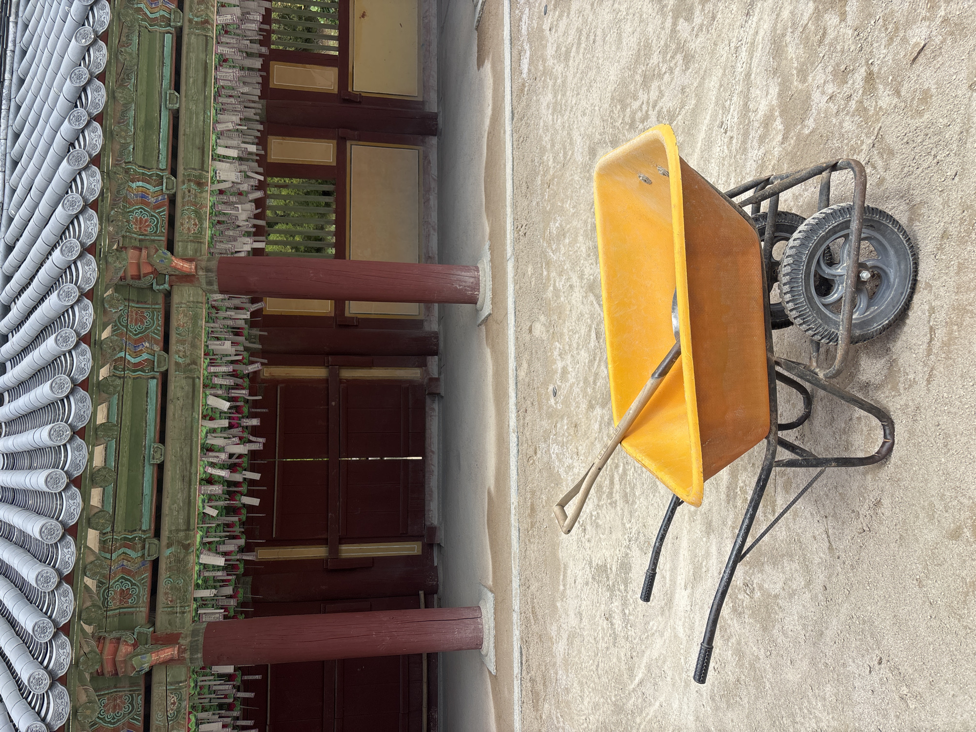 The image shows an orange wheelbarrow with a shovel inside, parked on a sandy ground in front of a traditional Korean building. The building features a tiled roof, ornate wooden eaves decorated with colorful paintings, and red pillars. Many small white papers are hanging from the eaves, possibly prayer tags or wishes. The building's walls are made of dark red wood, with some windows or openings revealing greenery behind them. The ground in the foreground is a mix of sand and dirt, with some patches of light gray concrete. The overall scene suggests an outdoor area, possibly a temple or historical site undergoing maintenance or renovation in Gyeongju-si, South Korea. There are no people or animals visible in the image.