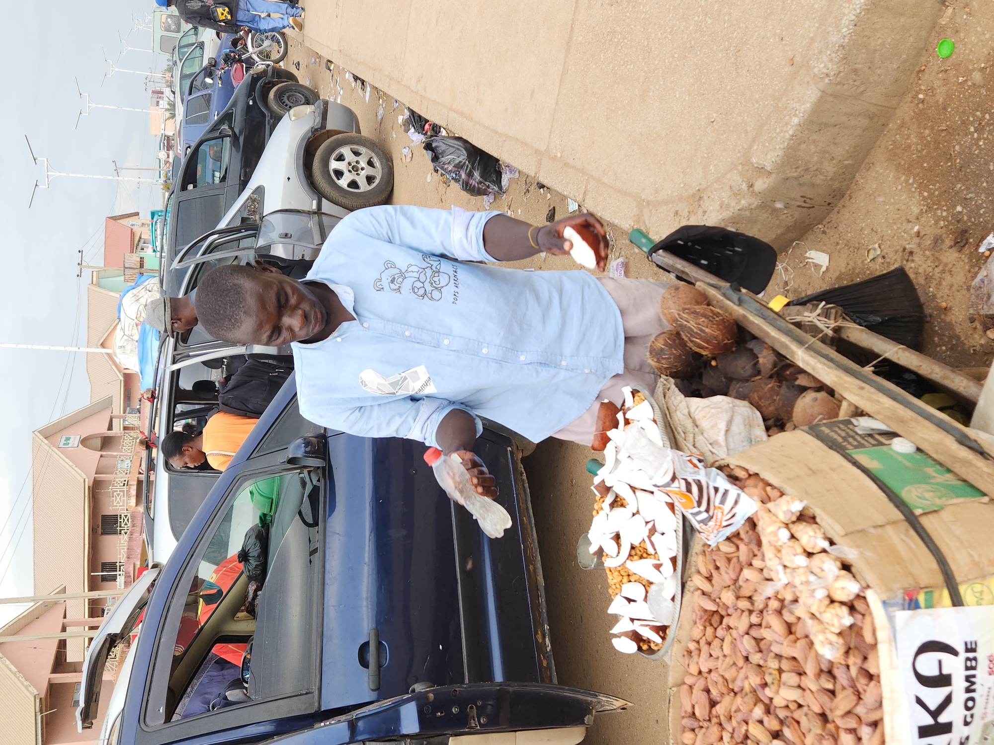 The scene shows a man standing on a street corner near a row of parked cars in Jos, Nigeria. He is selling coconuts, which are displayed in a tray with cut pieces and whole coconuts nearby. The man is holding a piece of coconut in one hand and a plastic bag in the other, possibly to serve a customer inside a dark blue car beside him. The roadside is littered with debris. The buildings across the street are pink.