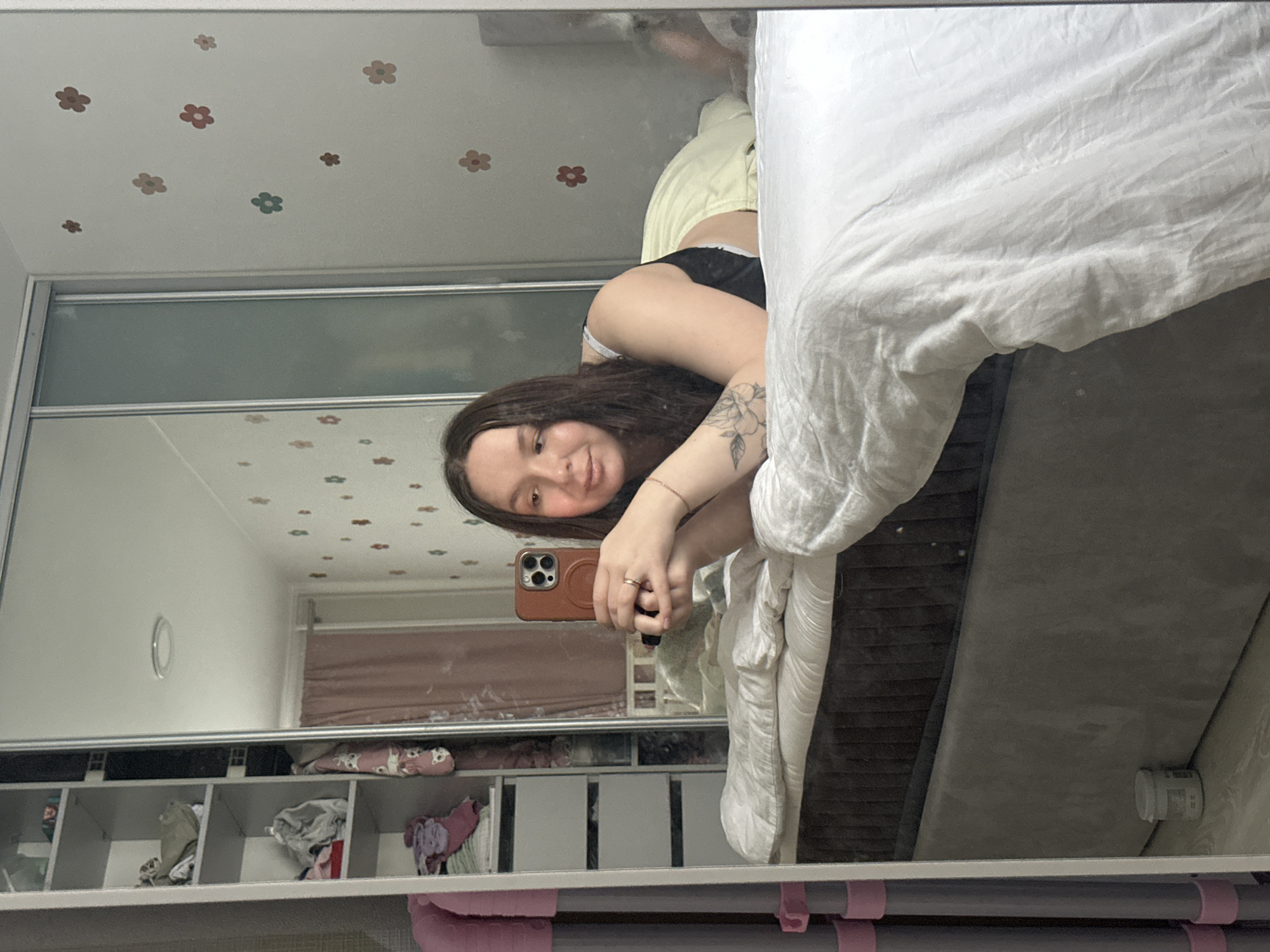 The scene depicts an interior view of a bedroom in Chaikovsky, Russia, captured through a mirror selfie. A female individual with long dark hair, wearing a black top and light yellow bottoms, lies prone on a bed with white crumpled bedding. She is propped on her forearms, holding a brown-cased smartphone (likely an iPhone) with both hands to capture her reflection. A tattoo of a floral design is visible on her left forearm, and she wears a ring on her left ring finger. Her expression is a slight smile directed at the camera.
The mirror appears to be part of a sliding door wardrobe on the left, which has open shelving stocked with clothes and other items. A frosted glass panel is part of the wardrobe. To the right, a light-colored wall features scattered small, multi-colored flower decals. A circular ceiling light is reflected overhead. In the reflection behind the camera's position, a light pink curtain is visible. The bed frame is upholstered in dark grey. On the floor to the left, a white lidded container rests next to the bed. Two vertical grey poles with pink caps are partially visible in the far left foreground. The flooring is a light wood laminate.
