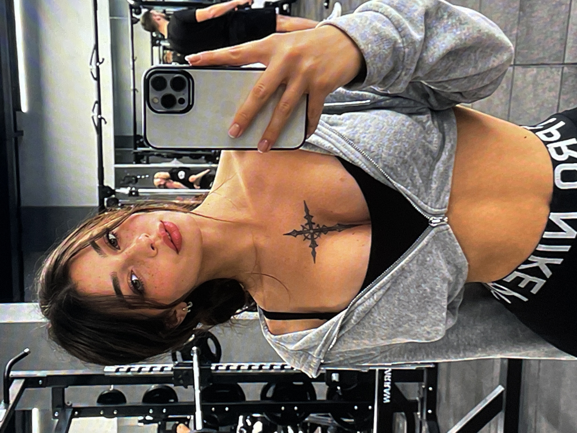 The image shows a young woman taking a selfie in a gym. She has dark hair, light skin with freckles, and is wearing a grey zip-up hoodie over a black sports bra and black leggings. A tattoo of a cross is visible on her chest. In the background, gym equipment, including weight racks and a squat rack, can be seen. Other gym-goers are also visible in the background. The location is identified as the city of Neubrandenburg, Germany, specifically within a gym facility.
