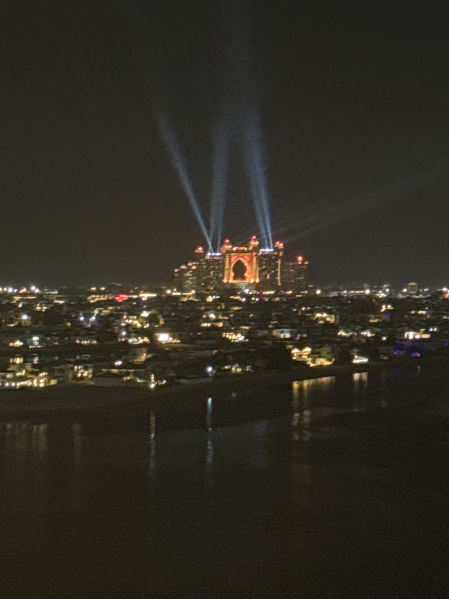 The image depicts a nighttime aerial view of a city illuminated by artificial lights. In the background, a prominent, ornate building with a large arched entrance and illuminated facade stands out, with beams of light shooting upwards into the dark sky. This landmark is identifiable as the Atlantis, The Palm hotel, located on the Palm Jumeirah island in Dubai, United Arab Emirates. The foreground shows a dark body of water, likely the Arabian Gulf, with reflections of the city lights. The cityscape is dense with buildings of varying sizes and illumination, suggesting a bustling urban environment. The overall impression is one of a vibrant, well-lit metropolis at night.