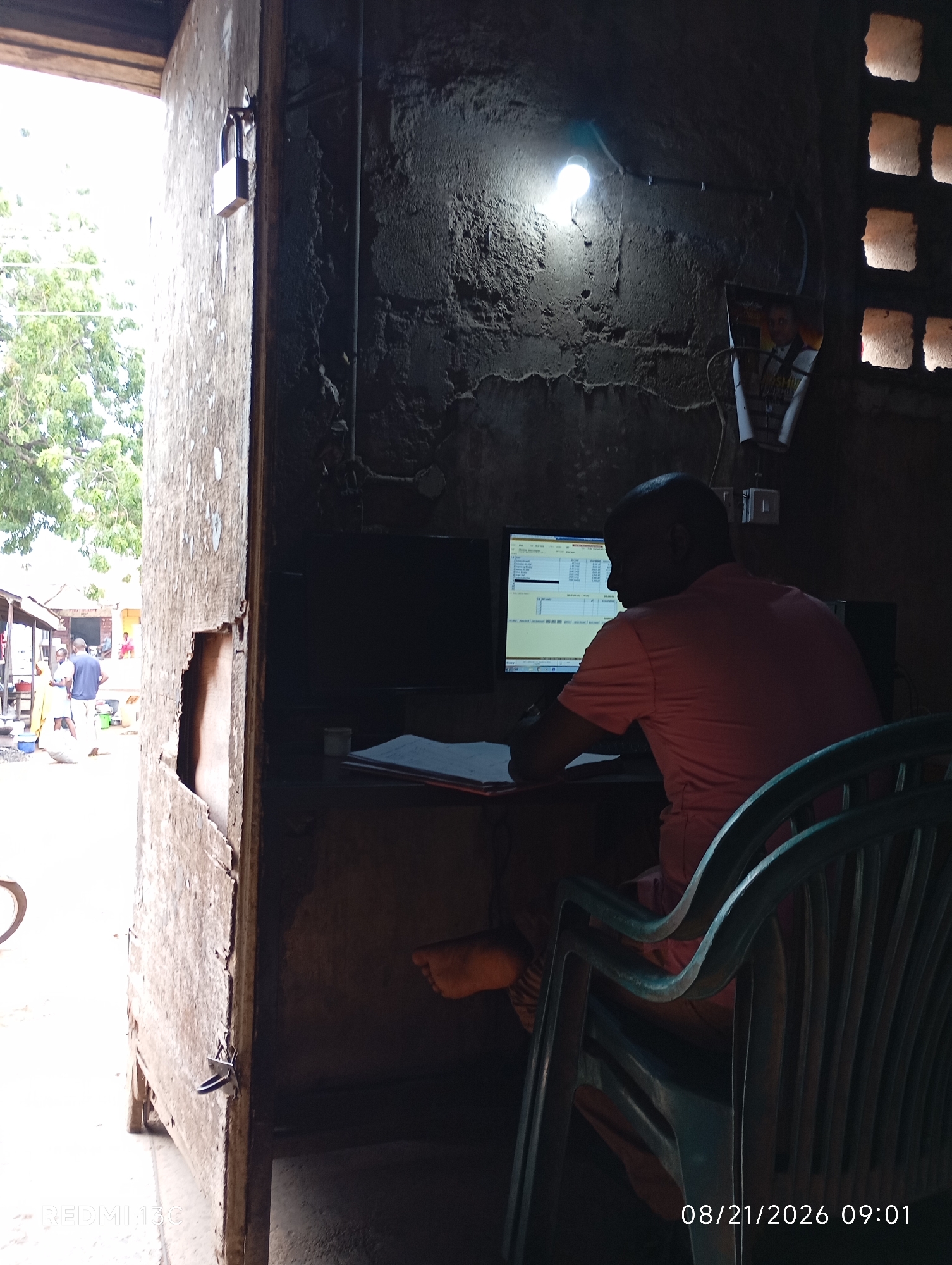 In Shendam, Nigeria, a man wearing a pink t-shirt is seated at a desk, facing a computer monitor. The room appears to be an interior space with rough, textured walls and a low-hanging light bulb illuminating the scene. A plastic chair is positioned in front of the desk. To the left of the interior space, an open doorway reveals an outdoor scene with trees and people in the distance. A portion of a wooden structure with a visible lock is on the left side of the image. The timestamp "08/21/2026 09:01" is present in the lower right corner.