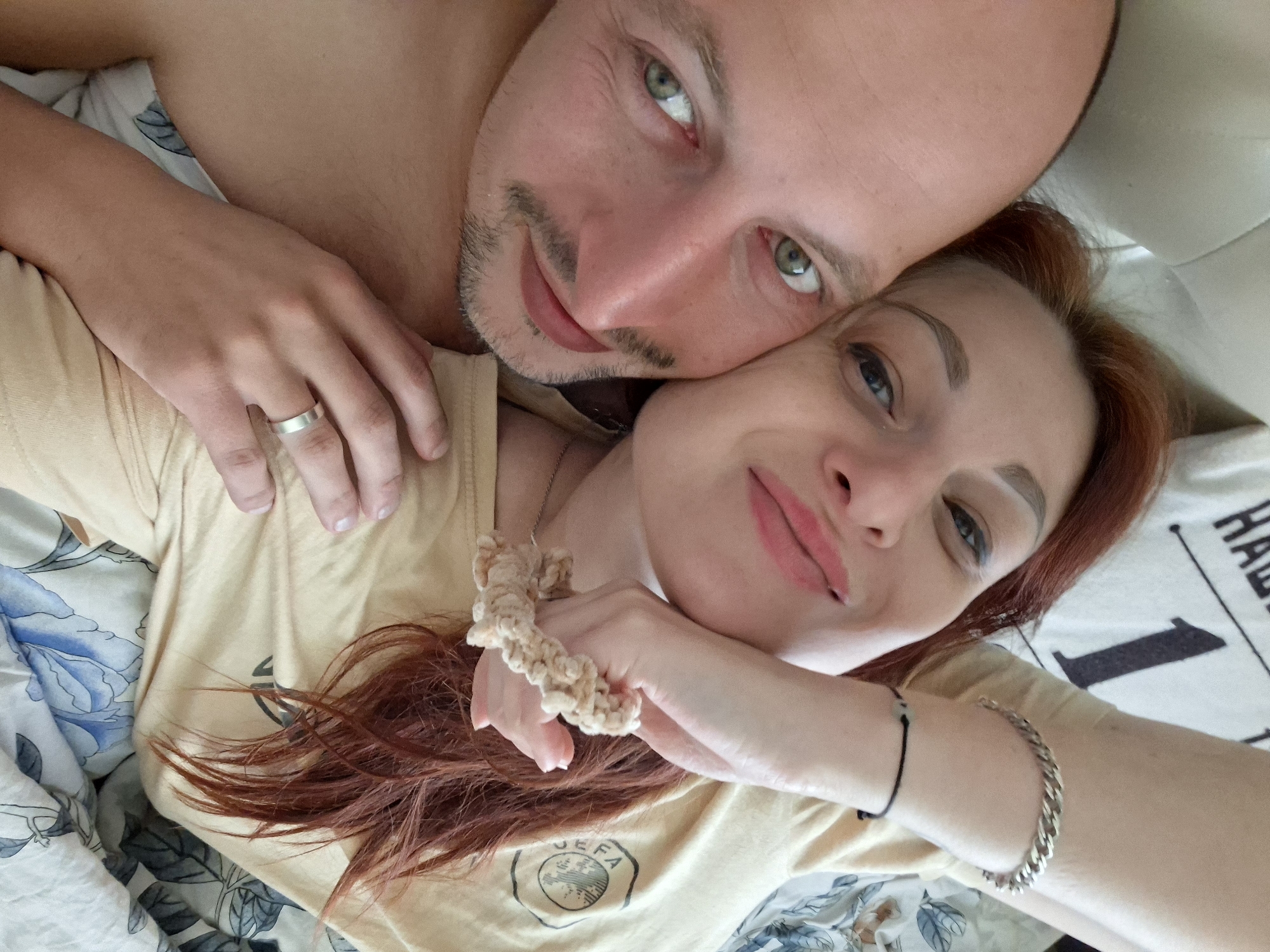 The image shows a close-up, overhead shot of a couple lying on a bed. The woman, on the left, has long, reddish-brown hair and is wearing a light yellow t-shirt. Her head is tilted, and her eyes are open, looking at the camera. Her left hand is resting on her face, and she's wearing a silver bracelet on her left wrist. A beige scrunchie is on her right wrist. The man, on the right, has short, dark hair and stubble. His eyes are also open and looking at the camera. He is wearing a silver ring on his left hand, which is resting on his chest. The couple is lying on a bed with floral patterned sheets and a white pillow. The background suggests an indoor setting, likely a bedroom. The image was taken in the city of Myadzyel, Belarus.