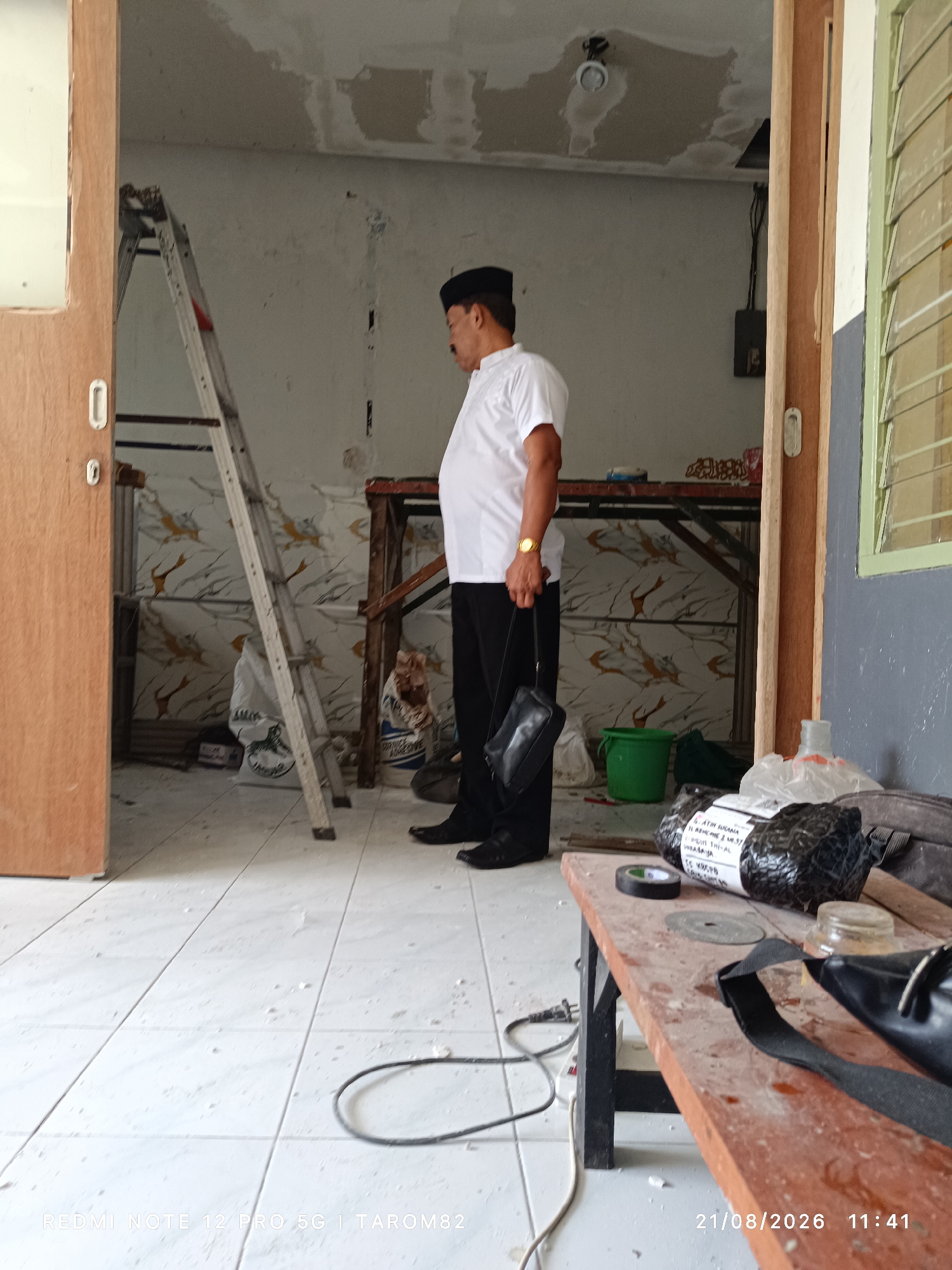 In Sruni, Indonesia, a man wearing a white shirt, black pants, and a black peci hat stands in a room undergoing renovation. He holds a black bag in his right hand. A silver ladder leans against a wall adorned with a patterned wallpaper. Various construction materials, including a green bucket, packaging, and a table, are visible. The floor is tiled, with scattered debris. A wooden door is on the left side of the image. A window with green shutters is on the right. The image is timestamped 21/08/2026 11:41.