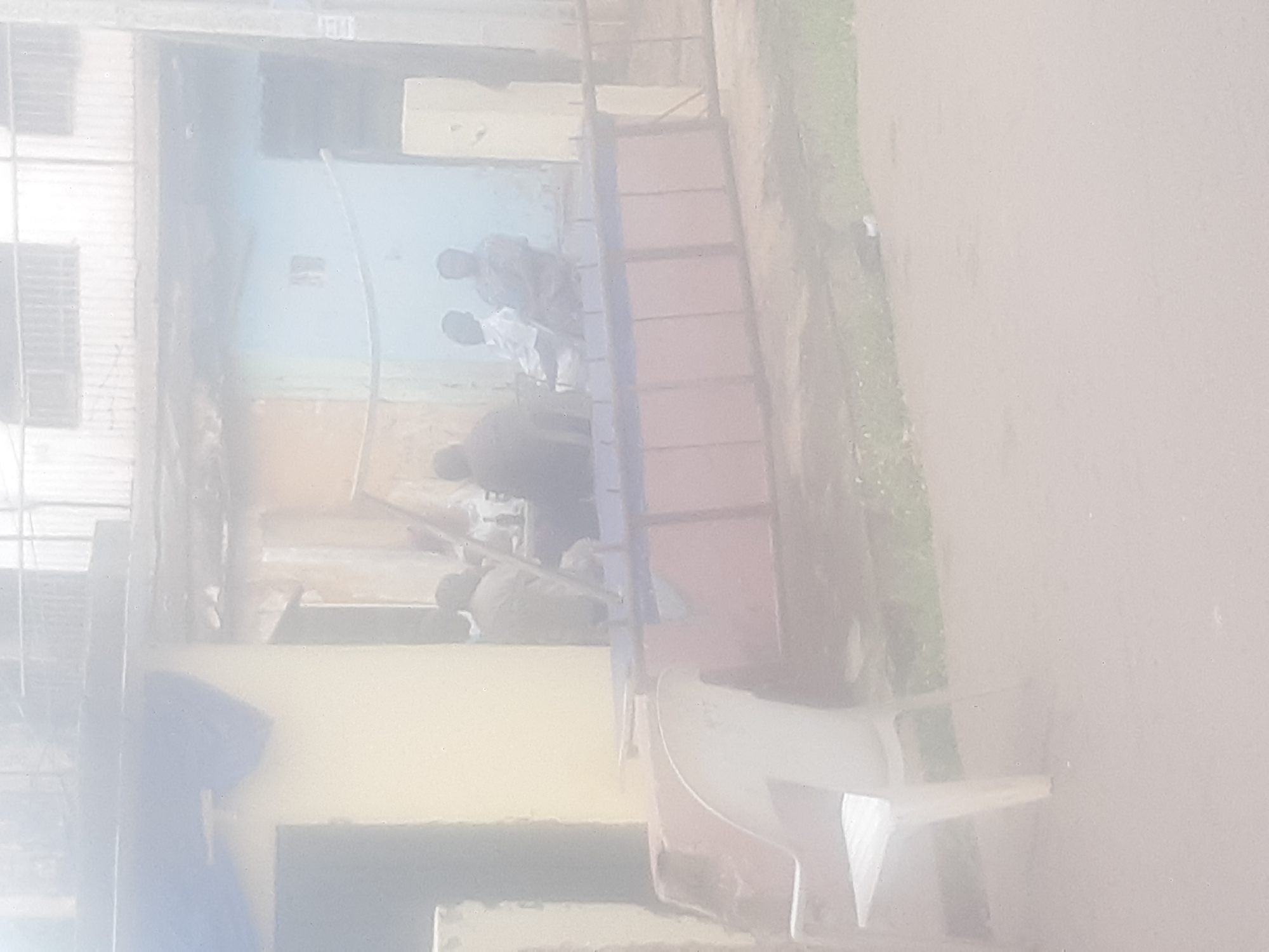 The image depicts a group of men sitting and conversing outside a building. The setting appears to be in a residential or commercial area, possibly a shopfront, with a light yellow and blue facade. A white plastic chair is prominently visible in the foreground. In the background, a multi-story building with windows can be seen. The overall atmosphere is casual, with the men engaged in conversation. The scene suggests a typical day in the city of Enugu, Nigeria, where such informal gatherings are common.