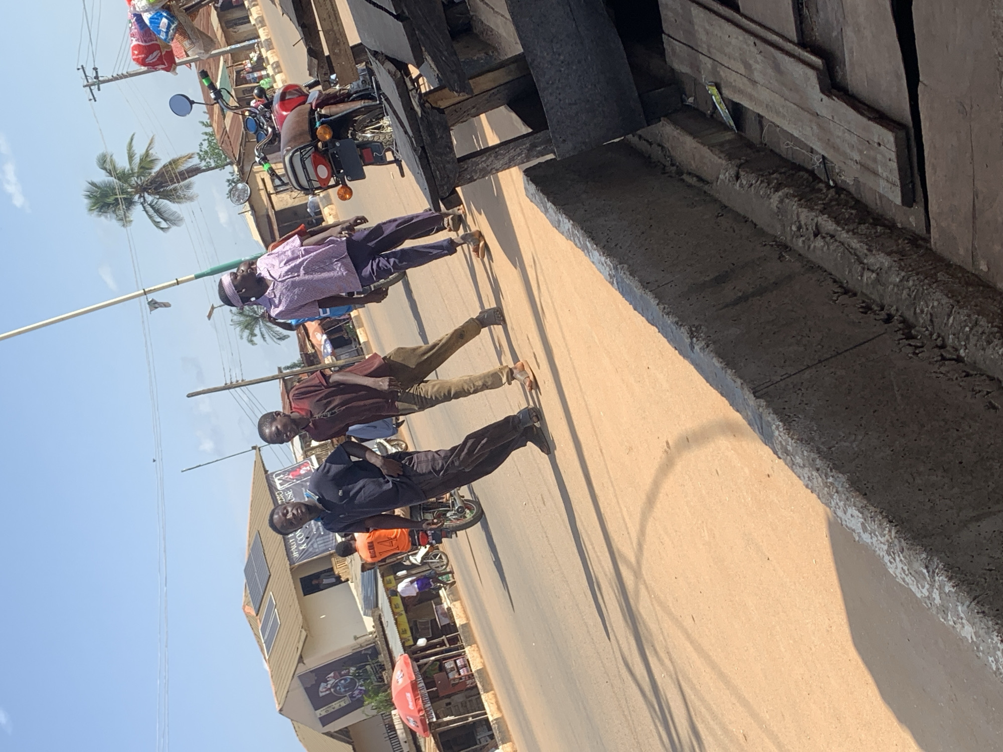 The image depicts a street scene in Kafanchan, Nigeria. Three young men are walking on a dirt road under a clear blue sky with scattered clouds. The man on the left wears a dark polo shirt and dark trousers. The middle man wears a brown collared shirt and khaki trousers. The man on the right is dressed in a pink and white checkered shirt and dark trousers, with a light-colored bandana on his head. In the background, buildings with tiled roofs, solar panels, and awnings line the street. A red umbrella marks a stall. A red motorcycle is parked to the right, with various goods displayed on a stall made of wood. A palm tree is visible in the distance. The ground is a light brown dirt. The overall scene appears to be a typical day on a street in a Nigerian town.