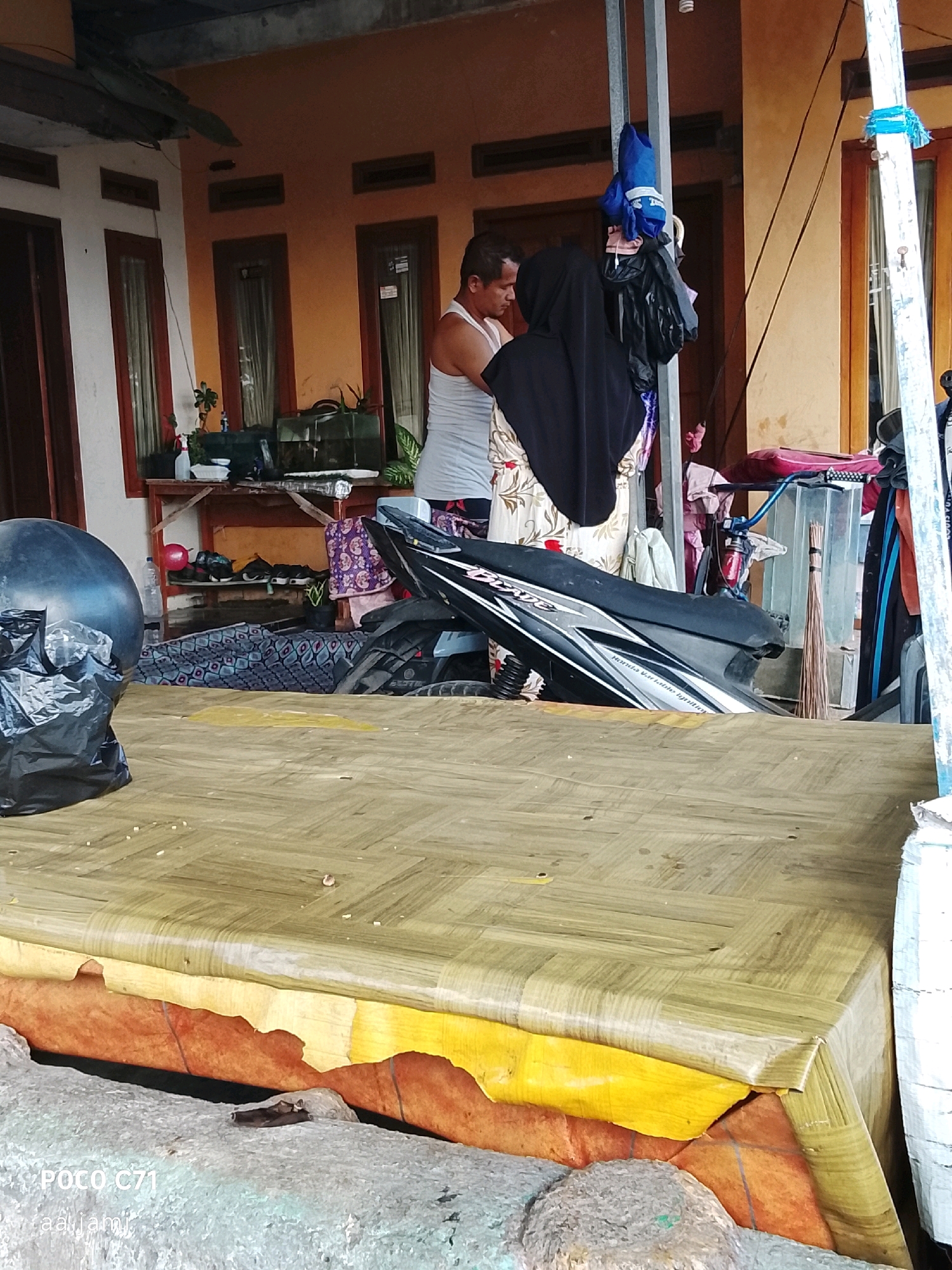 In front of a single-story house with an orange facade, a man wearing a white tank top and a woman wearing a black hijab stand near a motorcycle. The man appears to be looking down, possibly at something on the ground or in his hands. The woman is facing away from the camera, towards the man. A black motorcycle is parked in front of them, partially obscuring a table covered with a woven mat. To the left of the motorcycle, a large black garbage bag is visible. Behind the people and the motorcycle, a shelf unit holds various items, including potted plants and what appears to be a fish tank. Clothing is also seen hanging on a pole behind the individuals. The overall scene suggests a casual, everyday moment occurring in an outdoor residential area in Tasikmalaya, Indonesia.
