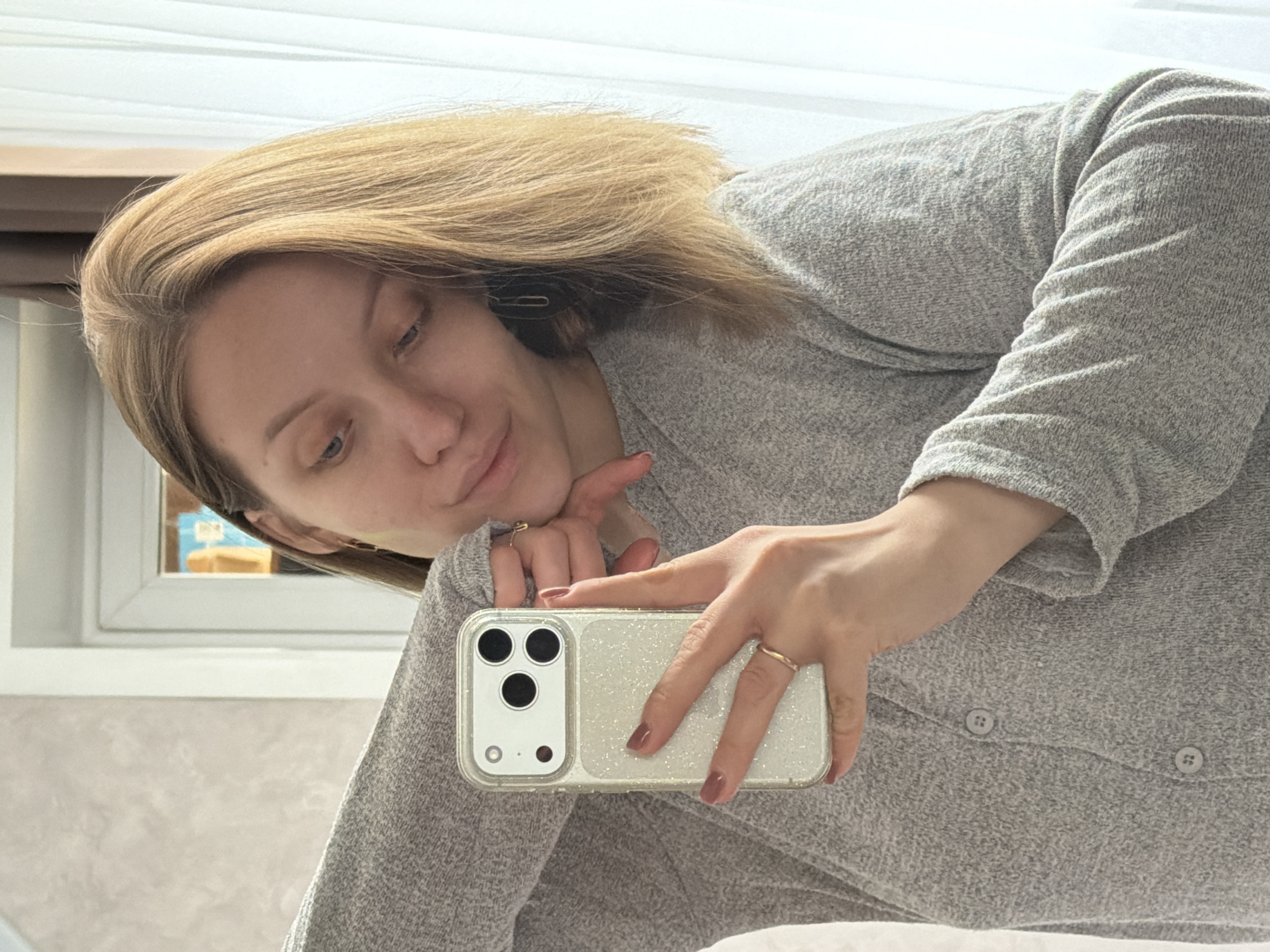 A woman with light brown hair is holding up a cell phone in a glittery case. She is wearing a light gray, long-sleeved, button-down shirt. Her hair is parted on the side, and she has a hairpin in her hair. She is wearing rings on both hands. The background includes a window with sheer white curtains, a light beige wall, and what appears to be a blurred view of the outside. There is no information in the image that confirms that it was taken in Reshetnikovo, Russia.