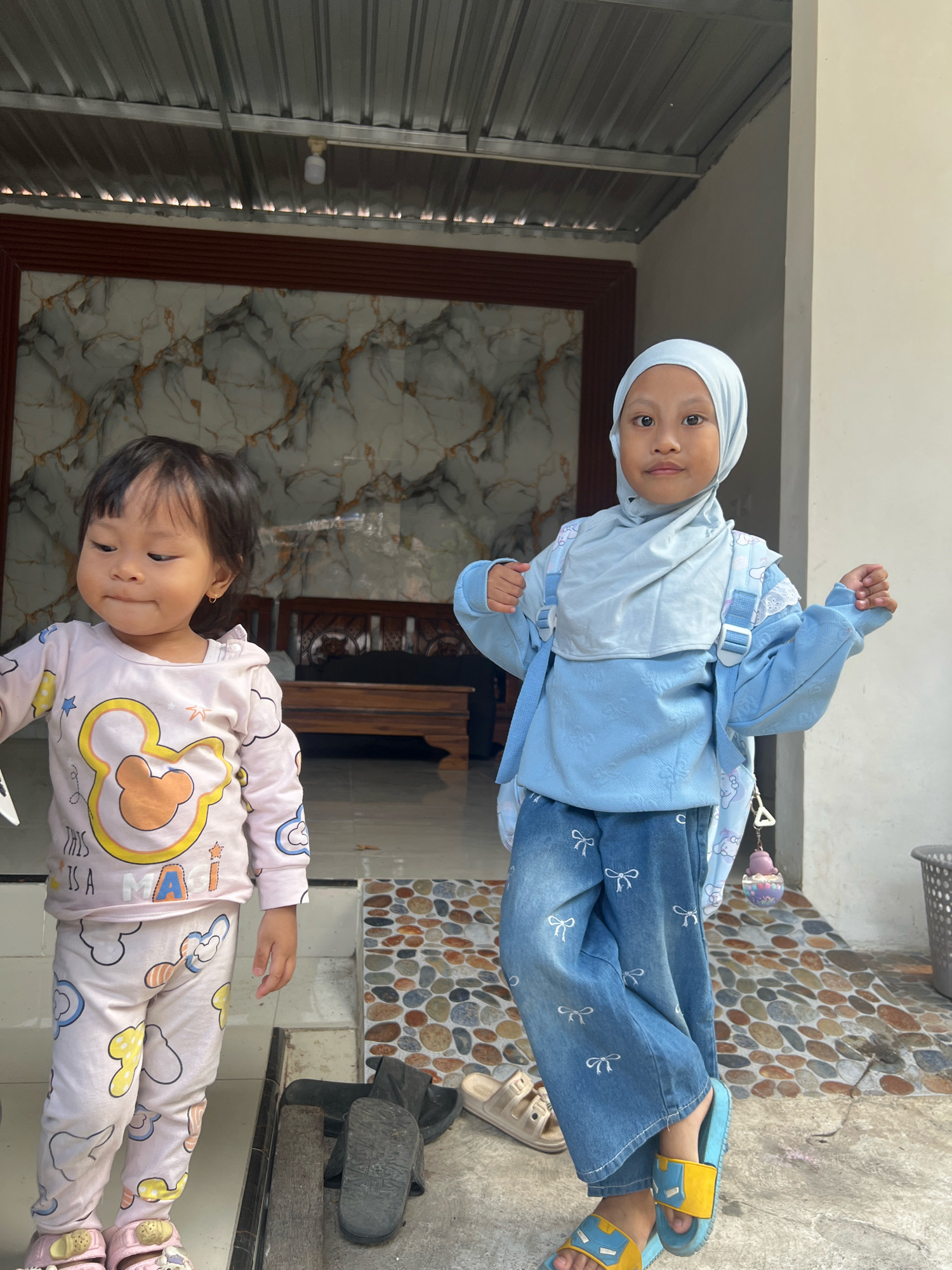 Two young girls stand outside a building in Kecamatan Pati, Indonesia. The girl on the left, with dark hair, wears a pink pajama set with a Mickey Mouse graphic. She is looking down and to her left. The girl on the right, wearing a light blue hijab and matching outfit with a backpack, is looking at the camera with her hands raised. She is wearing blue denim pants with a white bow pattern and yellow and blue sandals. They are standing on a tiled walkway in front of a building with a corrugated metal roof and a patterned wall. There is a wooden bench in the background.