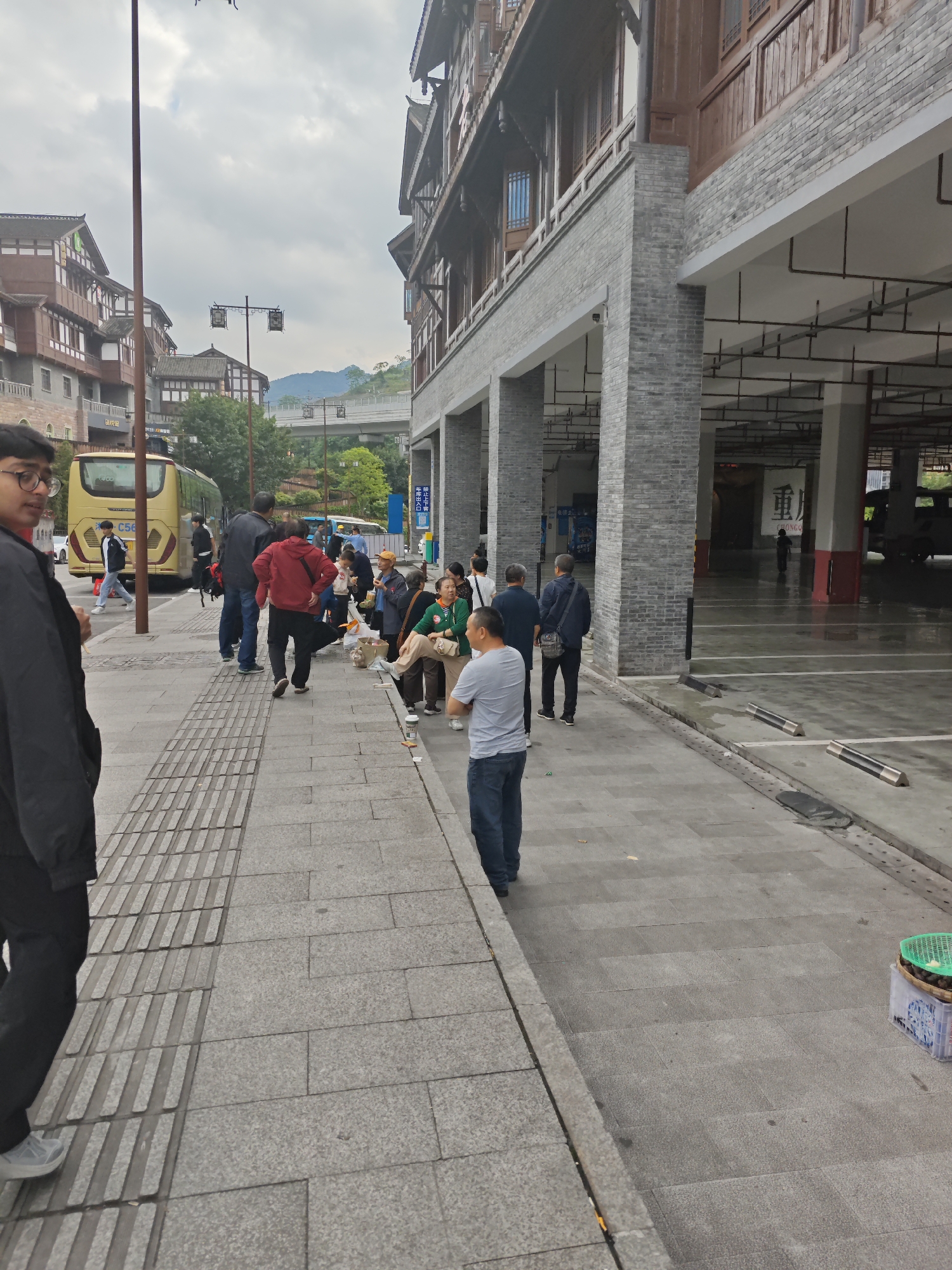 The image shows a street scene in Shapingba District, China, with pedestrians waiting near a bus. A yellow bus is parked along the street, and several individuals are standing or walking nearby. People of various ages are present, engaged in activities such as waiting, talking, or eating.
A modern building with a traditional-style facade, including gray brick columns and wooden architectural details, stands on the right side of the image. Underneath the building there is an open parking area, inside a figure is visible walking. The sky is overcast, suggesting a cloudy day. The foreground shows a paved sidewalk with tactile paving for the visually impaired. A container with what appears to be food or goods for sale rests on the sidewalk.