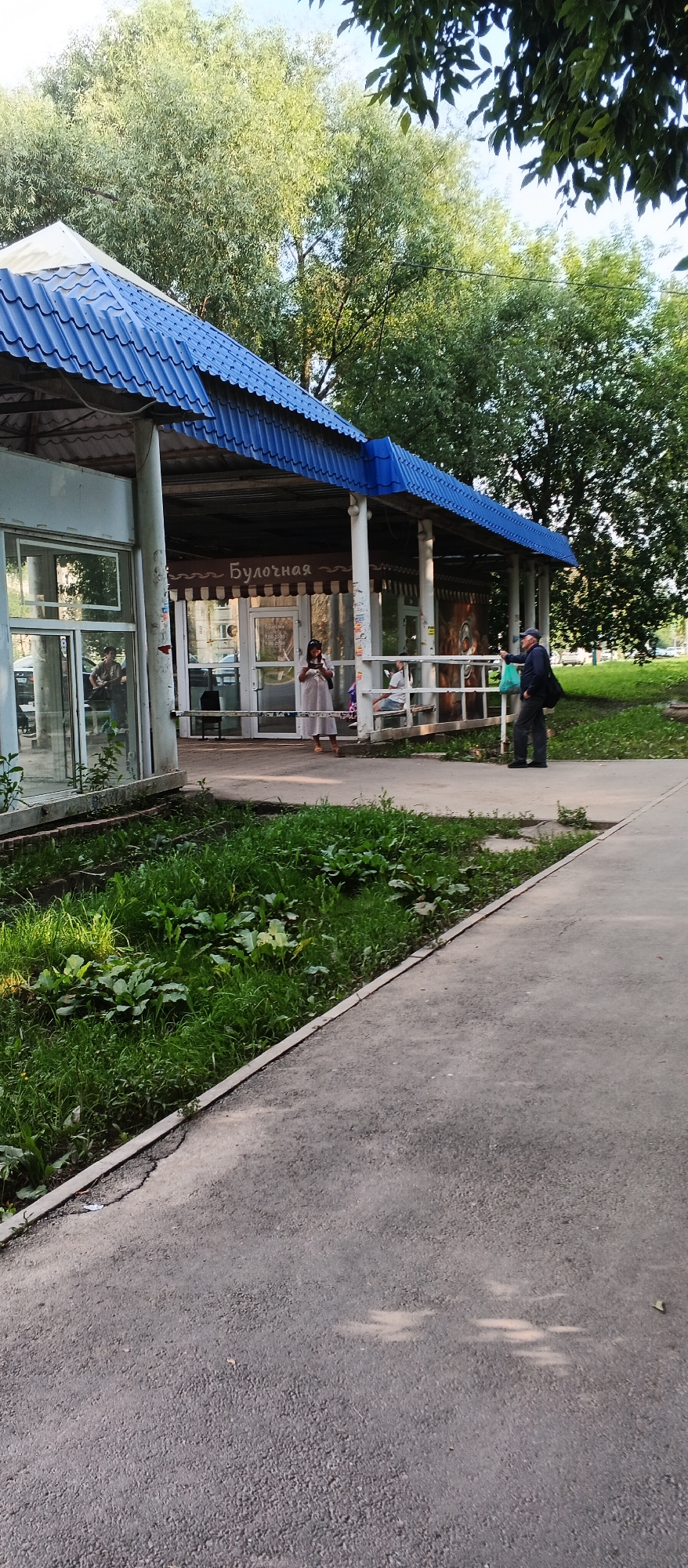 This image shows a small outdoor kiosk with a blue, corrugated metal roof. The kiosk has large glass windows and an open-air counter area. The word "Булочная" (Bakery) is visible above the counter. There are a few people present. A woman in a light-colored dress is standing at the counter. To her left, inside the kiosk, another person is partially visible. To the right, a man wearing a dark cap, dark jacket, and dark pants is standing outside, holding a green plastic bag and a dark bag over his shoulder. He appears to be interacting with someone inside the kiosk. In the foreground, there is a paved pathway and a strip of green grass and vegetation. Trees are visible in the background. The scene appears to be a typical street-side business in a city setting. Given the Cyrillic text, the location is likely in Russia.