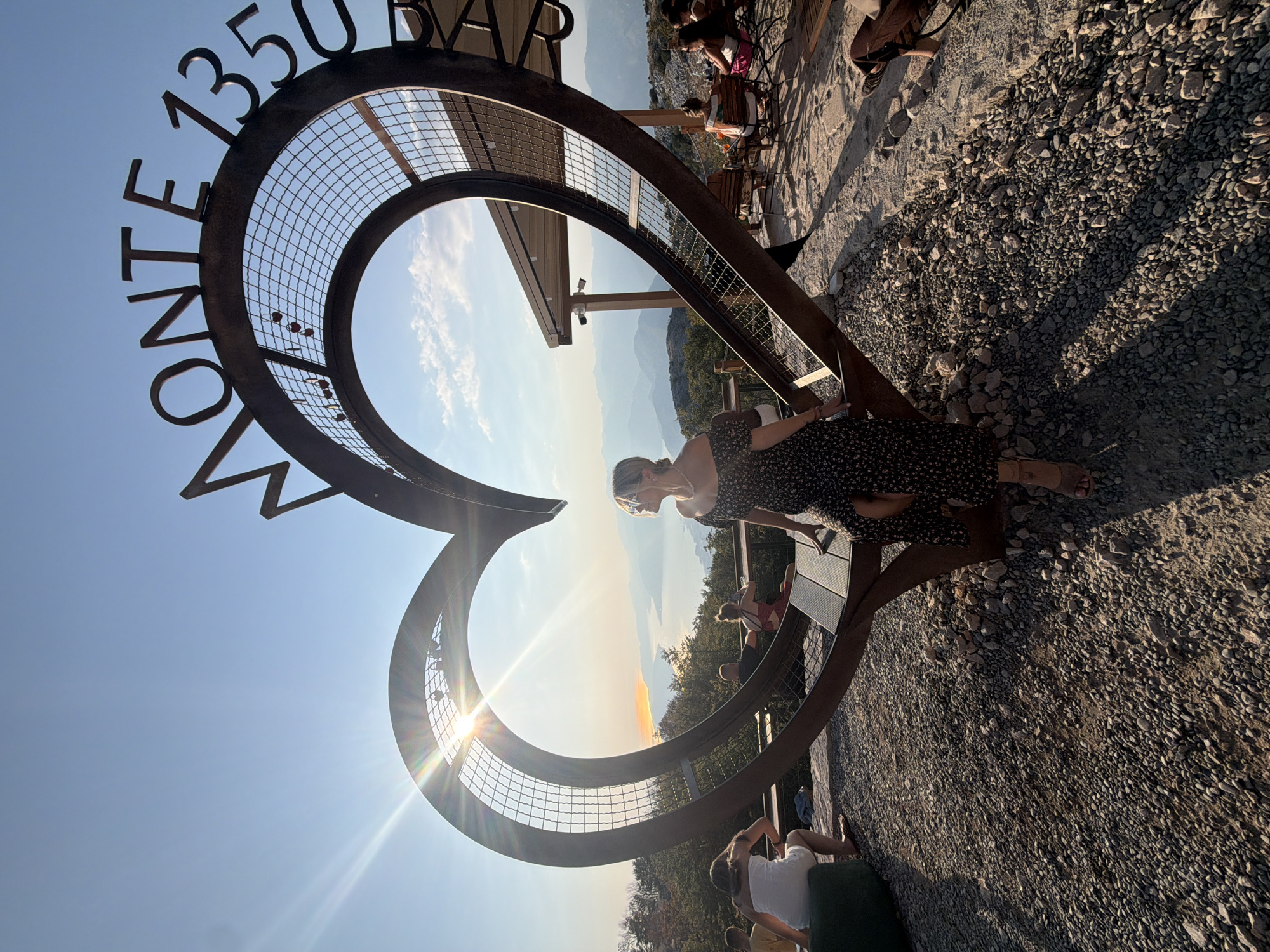 A woman in a dark floral dress sits on a metal bench inside a large heart-shaped sculpture with "MONTE 1350" inscribed on its upper arc. The sculpture is positioned outdoors on a rocky, gravelly surface. The background shows a scenic view of a body of water surrounded by distant mountains under a clear blue sky with the sun shining brightly. Several other people are visible in the background, some seated at tables near a covered structure. This sculpture is located at Monte 1350 in Kuk, Montenegro, a viewpoint offering panoramic vistas of the surrounding landscape.