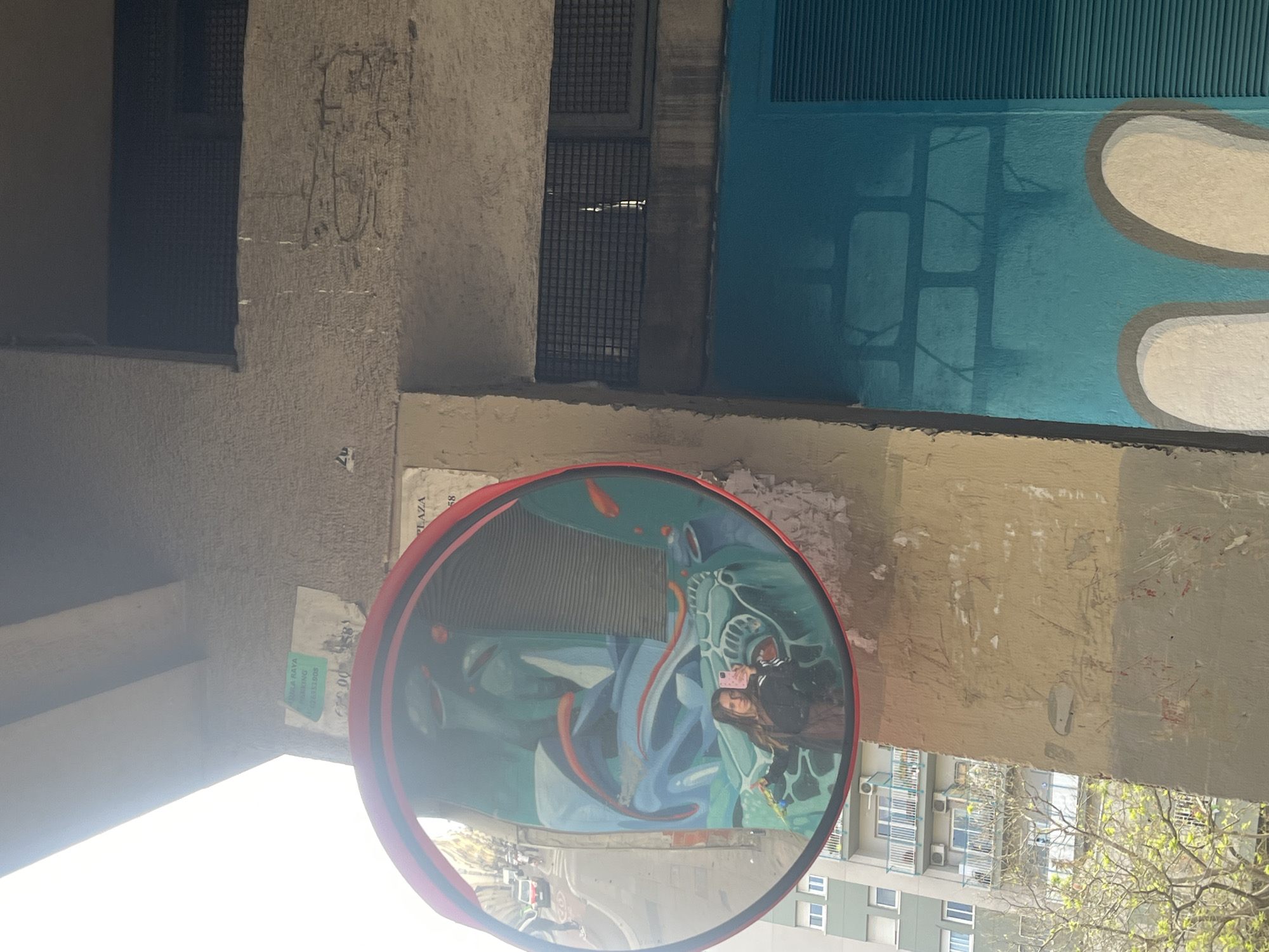 A vibrant street scene depicts an urban environment, likely in Barcelona, Spain. A large, red-rimmed convex traffic mirror is centrally positioned on a textured beige wall, reflecting a detailed view of its surroundings. Inside the mirror, a young woman with long dark hair, wearing a black jacket, holds a pink smartphone to take a photograph, capturing her reflection and the street art behind her.
The mural reflected in the mirror features a complex design with abstract blue and green shapes, a stylized face, and several elongated orange forms resembling fish. Directly visible on the wall adjacent to the mirror is another section of street art, a bright blue surface adorned with large, smooth white and grey abstract shapes.
The overall scene is characterized by concrete architecture, including plain walls, grated windows, and distant buildings with visible air conditioning units reflected in the mirror. The lighting suggests it is daytime with clear conditions, casting some shadows.
Visible text on papers taped to the wall near the mirror includes "LA PLAZA," "PZA. DEL...", and a green sticker stating "PLAZA GAYA PARKING" along with the phone number "933131308". Faint, indistinct graffiti can also be seen on one of the concrete walls.