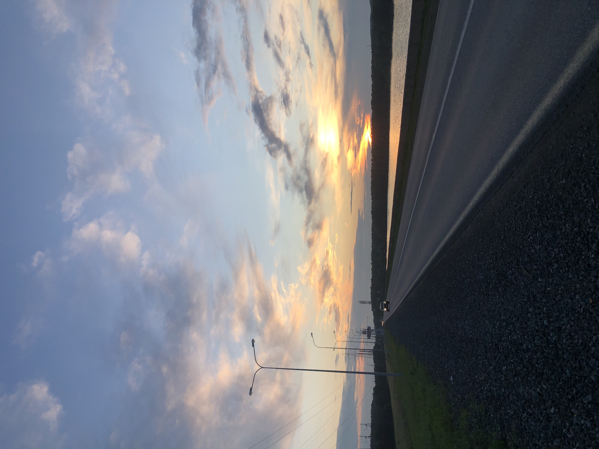 The image depicts a road in the city of Chaikovsky, Russia, likely near the Kama River, at sunset. The sky is filled with dramatic clouds in shades of blue, white, and orange, with the sun setting on the horizon. The road, paved and with a white line marking the lane, stretches into the distance, bordered by a gravel shoulder on the left and a body of water on the right. A dark green tree line forms the far bank of the water. Several streetlights line the left side of the road, with power lines and transmission towers visible further in the background. A single car, with its headlights on, is traveling on the road in the distance. The overall scene conveys a sense of tranquil evening travel.