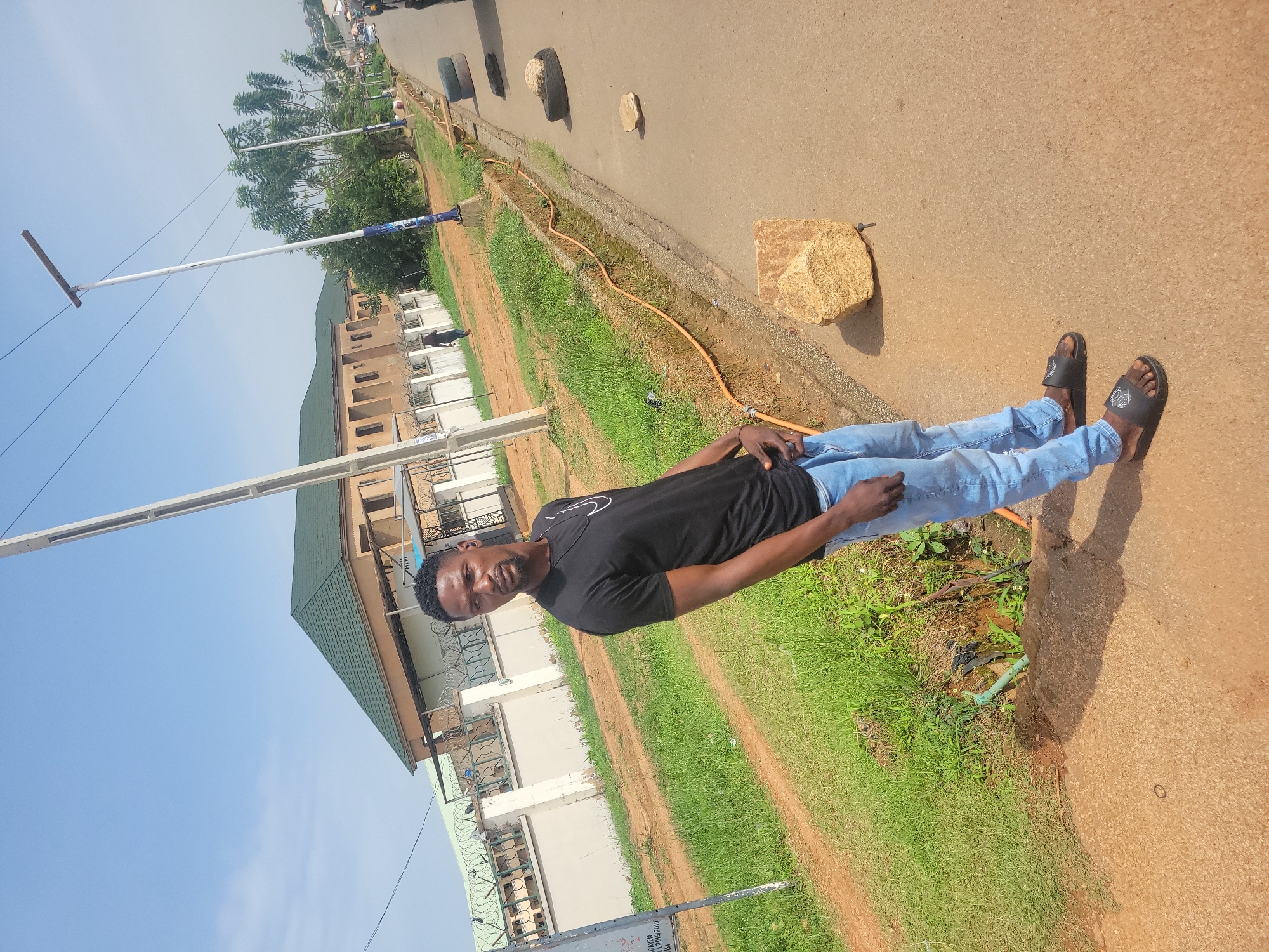 A full-body, outdoor, eye-level, medium shot captures a young Black male standing on a paved road. He is facing slightly to his right, looking over his left shoulder at the camera. He has short black hair and a goatee, and is wearing a black t-shirt with a white logo on the left chest, light blue jeans, and black slide sandals. His left hand is in his front jeans pocket, and his right arm is bent with his hand resting on his waist.
The background features a beige building with a green roof, surrounded by a white fence topped with barbed wire. To the left of the building is a grassy area with a dirt path. The road the man stands on is paved and has a drainage ditch running alongside it, containing an orange pipe. Several tires and large rocks are scattered along the side of the road. In the distance, trees and more buildings are visible under a clear blue sky. The scene appears to be in an urban or suburban area.