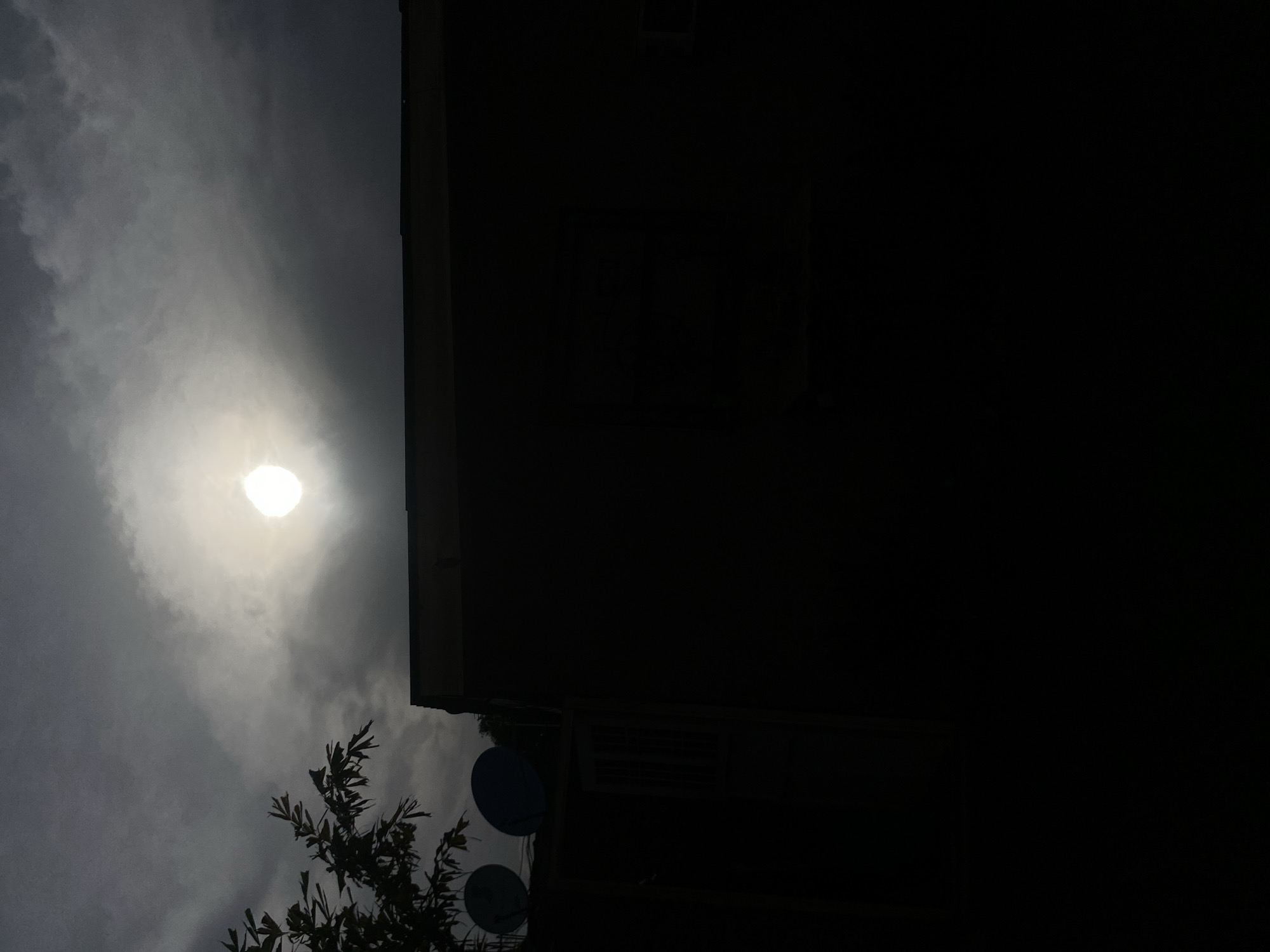 The image appears to be a low-angle shot, possibly taken from the ground, depicting a portion of a dark building silhouette against a cloudy sky with the sun obscured but still visible through the clouds. A tree branch with leaves is partially visible in the lower left corner. On the building, two satellite dishes are discernible. The location of the image is stated to be Barden Yana, Nigeria.