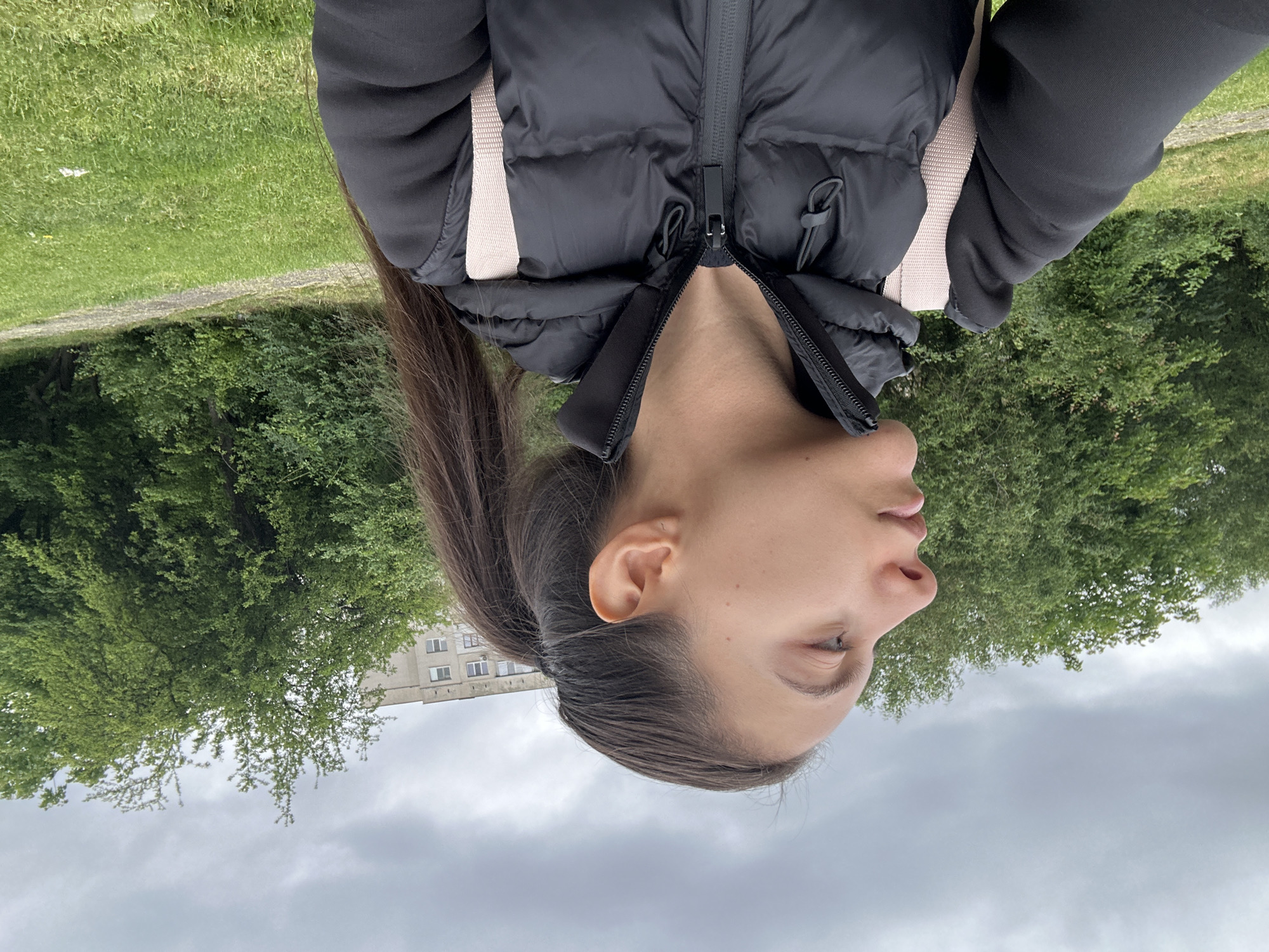 The image is a low-angle, slightly off-center photograph of a woman wearing a black puffer vest with a cream-colored strap, seemingly taken outdoors in Sofia, Bulgaria. The woman has dark hair pulled back in a ponytail, and her face is in profile. In the background, there are green trees, a grassy area, a building with multiple windows, and a cloudy sky. The photograph is oriented upside down.