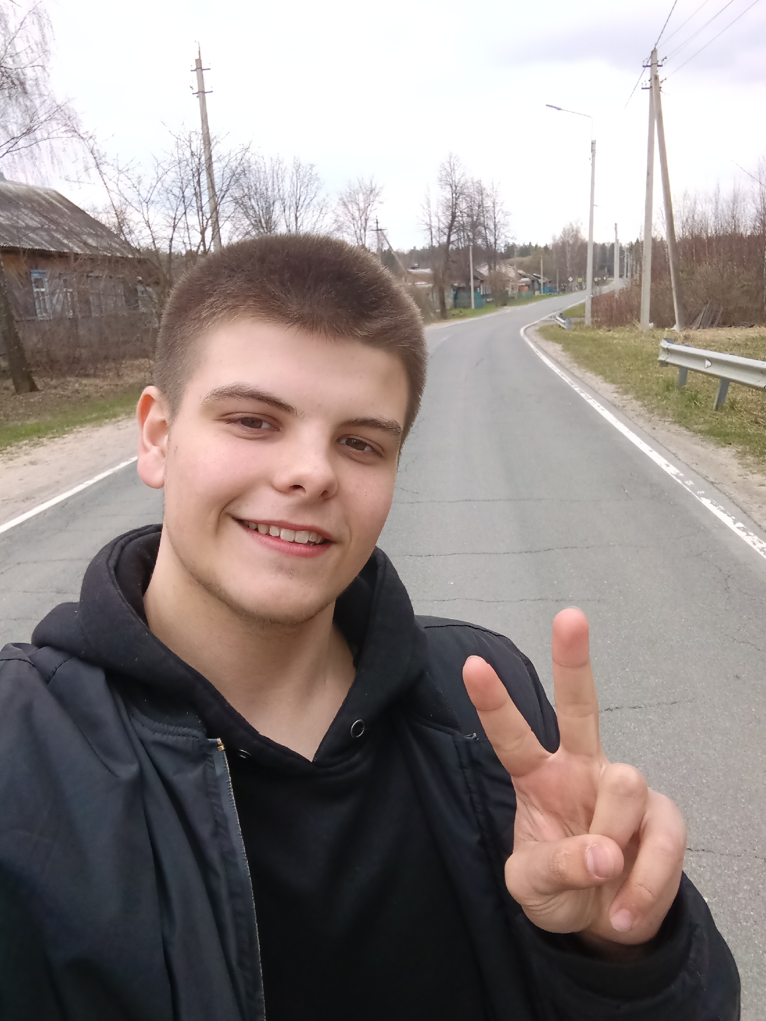 A young man is taking a selfie on a paved road in Красный Маяк, Russia. He is smiling and holding up a peace sign. He is wearing a black hoodie under a black jacket. The road stretches into the distance with utility poles along the sides. There are houses visible on either side of the road, and the sky is overcast. A metal guardrail is on the right side of the road.