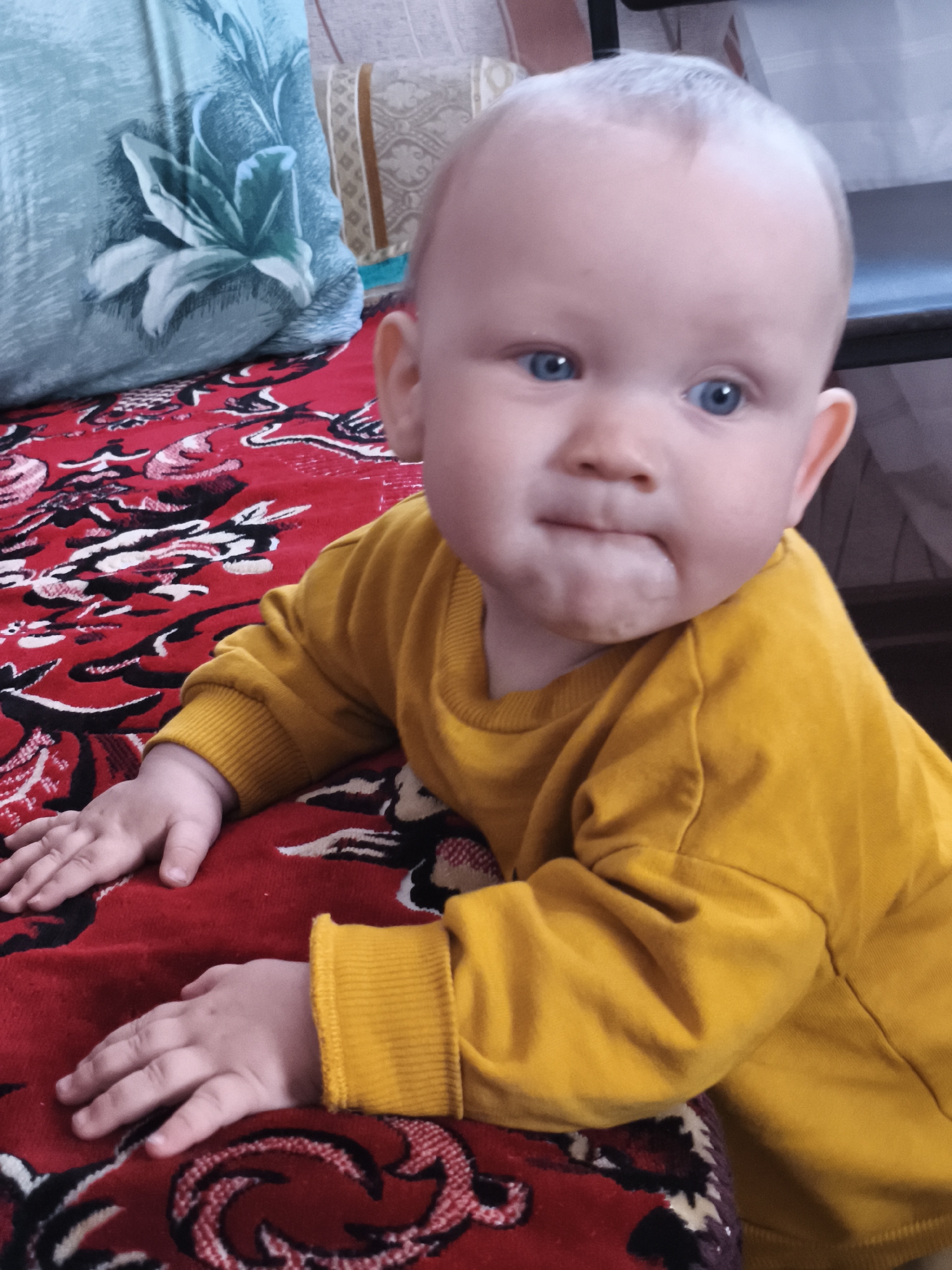 A baby with blue eyes and light hair, wearing a yellow sweatshirt, is lying on a red blanket with a floral pattern. The baby is positioned on the left side of the frame, facing towards the viewer with their head tilted slightly upwards. Their hands are resting on the blanket. In the background, to the left, is a light blue pillow with a white floral design. To the right of the baby's head, a portion of a beige and brown striped pillow is visible. The setting appears to be indoors, likely in a domestic environment. The location is identified as the city of Chaikovsky, Russia.