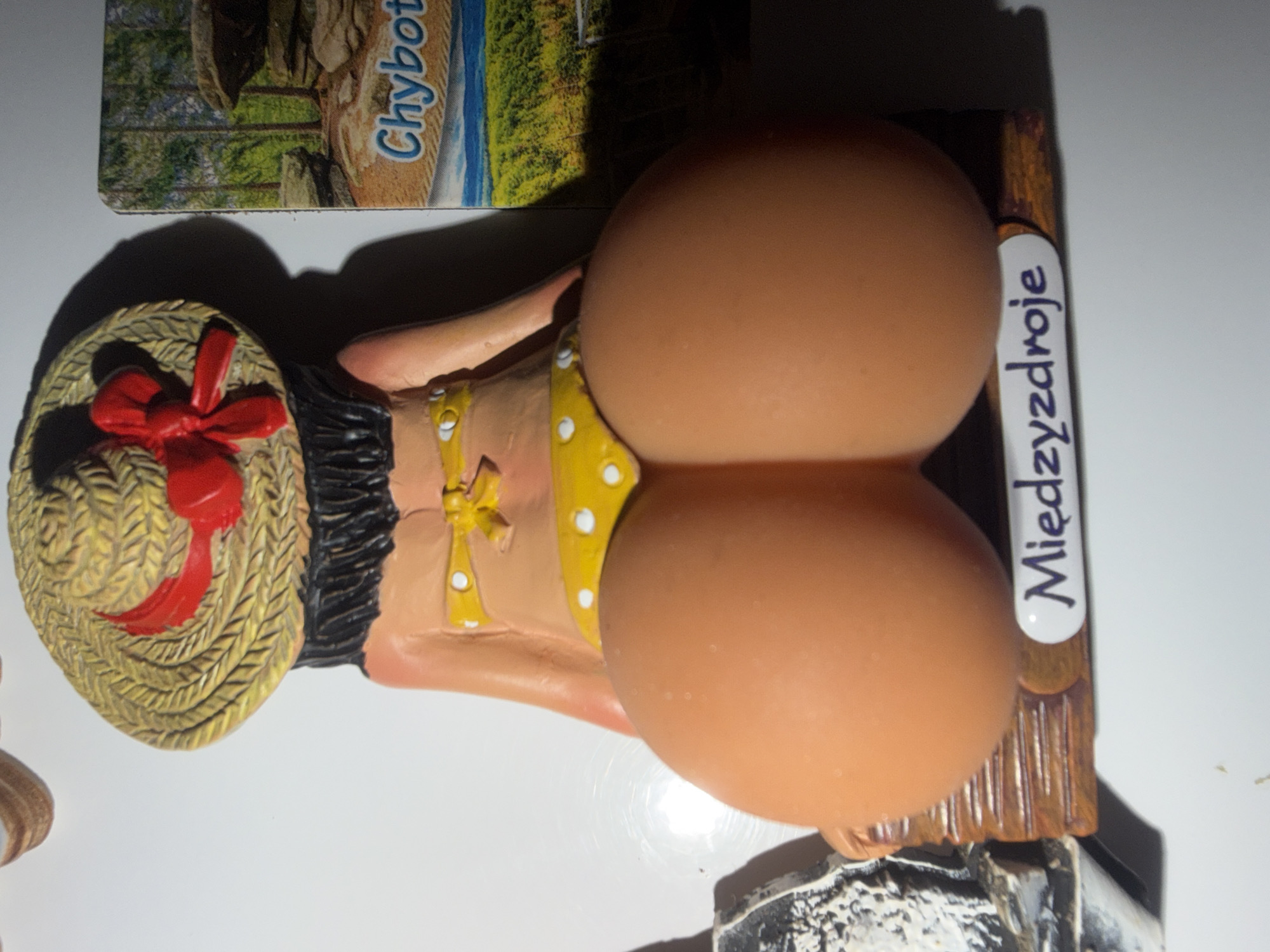 The image displays a souvenir magnet shaped like a woman's rear end wearing a yellow polka dot bikini and a straw hat with a red ribbon. The souvenir is placed on a white surface, with a postcard of a forest landscape featuring large rocks and the text "Chybot" visible to the right. Below the souvenir's base, the word "Miedzyzdroje" is written. The souvenir does not appear to be located in the city of Szczecin, Poland, but rather in a place associated with the text "Miedzyzdroje".