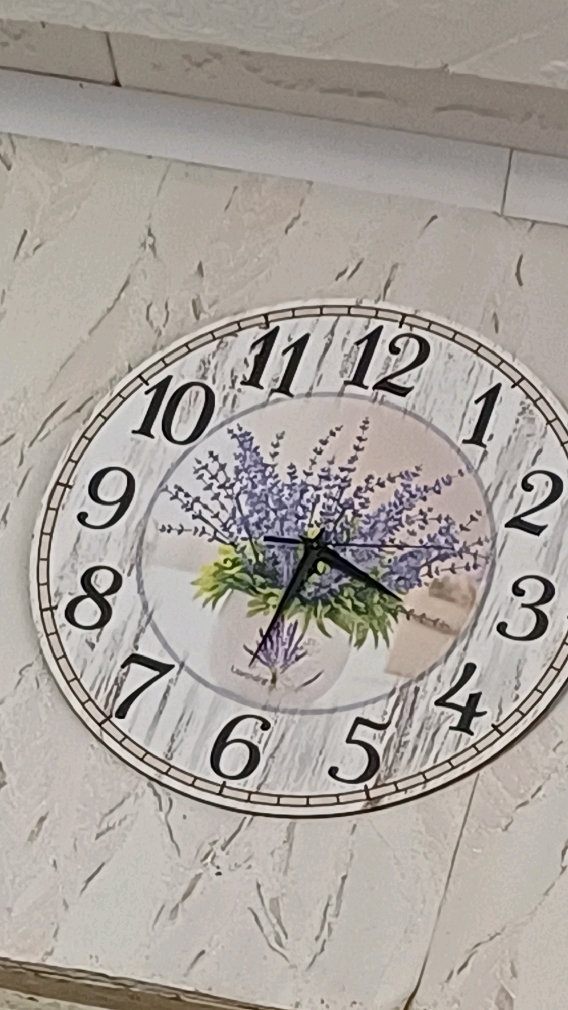 The image shows a close-up of a round wall clock with a decorative floral design of lavender in a pot. The clock face has black Arabic numerals indicating the hours. The clock hands are black. The clock is mounted on a textured, off-white wall, which appears to be made of plaster or a similar material. Above the clock, a white painted pipe or conduit runs horizontally along the ceiling or upper wall. The overall impression is of a domestic or interior setting. There is no indication of the city of Stavrovo, Russia in the image.