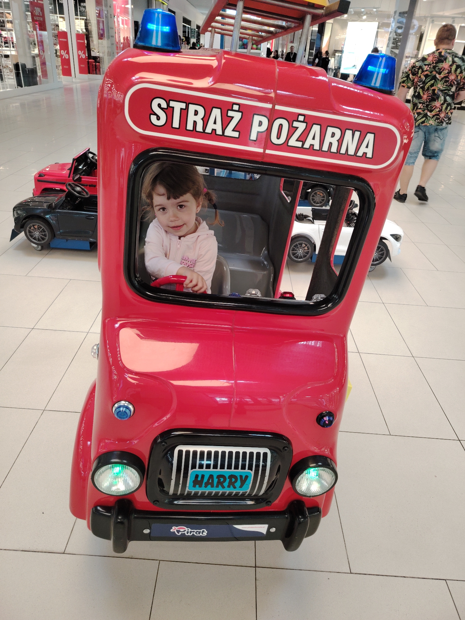 A young girl with dark hair in pigtails sits in the driver's seat of a bright red, toy fire truck. The fire truck has the words "STRAŻ POŻARNA" written on its front. The girl is wearing a light pink hooded jacket. In the background, other toy cars are visible, including a black jeep and a white car. The scene appears to be indoors, likely in a shopping mall, with tiled flooring and shop windows visible in the distance. A man wearing a floral shirt and denim shorts walks away from the camera in the background. The fire truck has blue flashing lights on its roof and a green headlight. A license plate with the name "HARRY" is visible on the front of the truck. A sticker that reads "Pirate" is on the lower front bumper. The image is taken at a slight angle, with the toy fire truck being the primary subject. The location appears to be a shopping center in Wołomin, Poland, given the context provided, although this cannot be confirmed solely from the visual information.