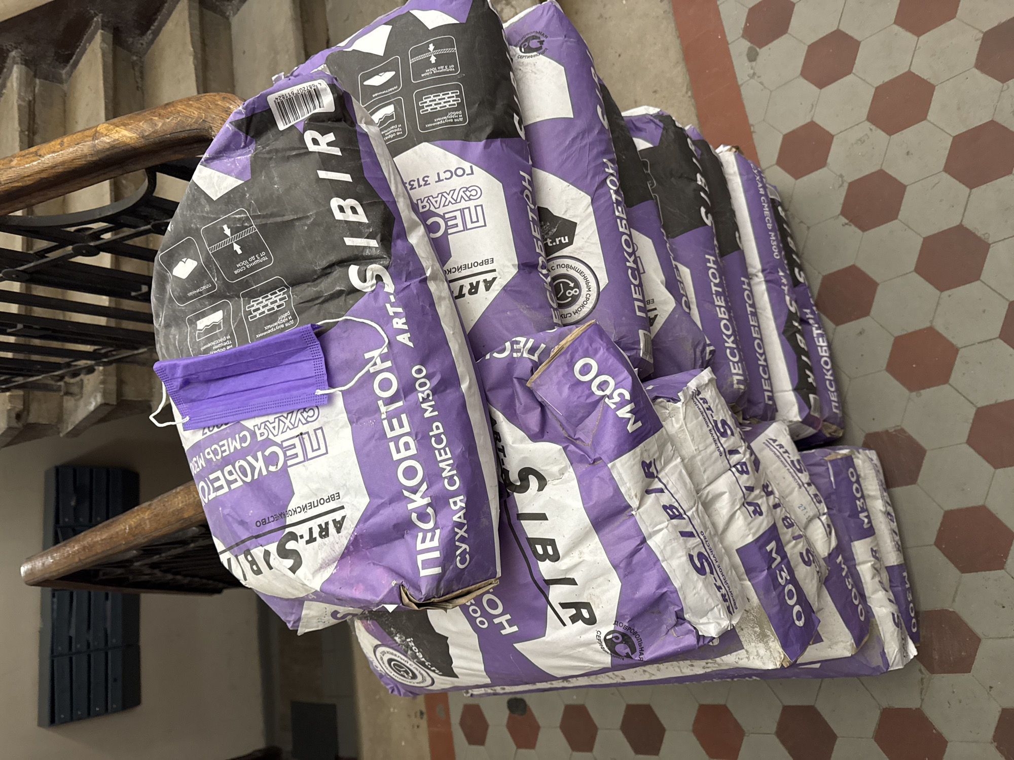 The image shows a stack of approximately nine bags of "Пескобетон М300" (Pesсobeton M300) dry sand-concrete mix, a common construction material, resting on a floor with a hexagonal tile pattern. The bags are purple and white, with "ART-SIBIR" prominently displayed. A discarded purple medical mask rests atop one of the bags.
The floor is tiled in a pattern of gray and red-brown hexagons. The stack of bags is near a wooden handrail and a wall with what appears to be a dark-colored cabinet or mailbox. Based on the Cyrillic text and the style of construction material, the location is likely Moscow, Russia.