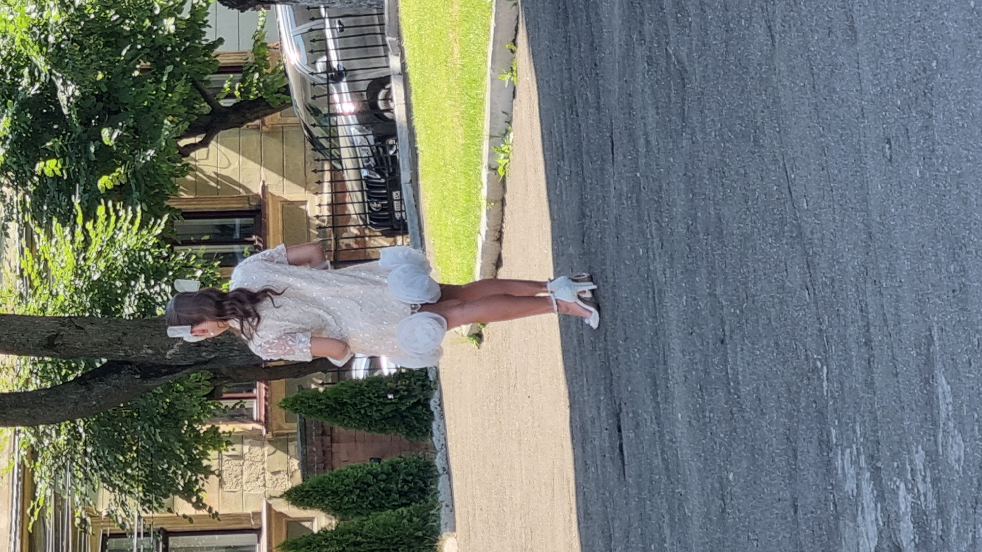 A young woman, seen from behind, walks on a paved surface. She is wearing a short, light-colored dress with short sleeves and a decorative hem that resembles fluffy white flowers. Her hair is dark and pulled back, with a white accessory at the crown. She is also wearing white high-heeled shoes with ankle straps.
The background consists of a building with stone walls and windows, a tree with green foliage, and a black metal fence. Beyond the fence, a dark-colored vehicle is visible parked on a grassy area. The scene appears to be outdoors in a city setting. The overall lighting suggests it is daytime.
Based on the architectural style of the building with ornate details and the surrounding environment, this scene is likely located in Chernivtsi, Ukraine.