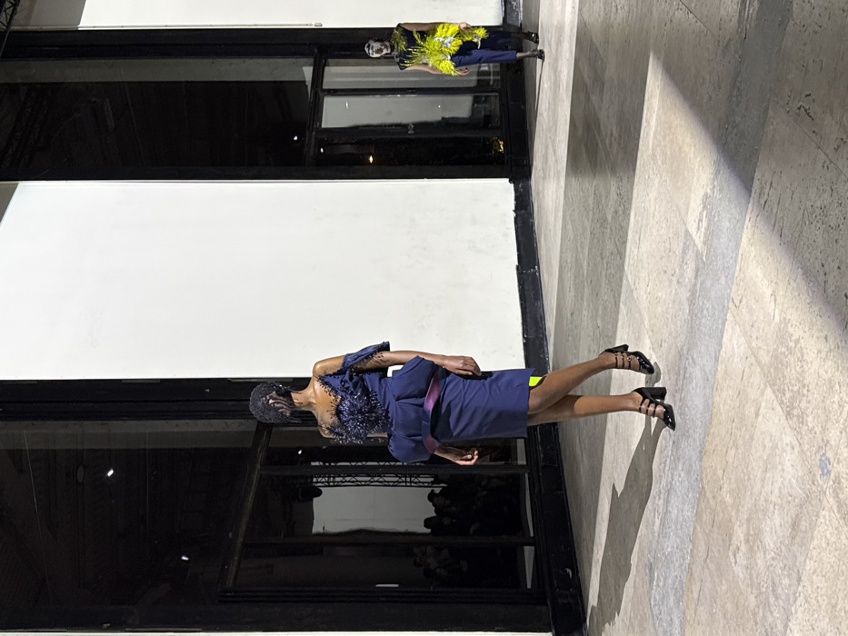 Two models stand beside a white wall with black framed windows. The first model is in a yellow shirt and dark pants. The second model wears a navy dress with an ornate navy shoulder piece, a pink sash, and black heels. The floor is a light, neutral color, marked with light-colored squares. An audience is visible through the window.