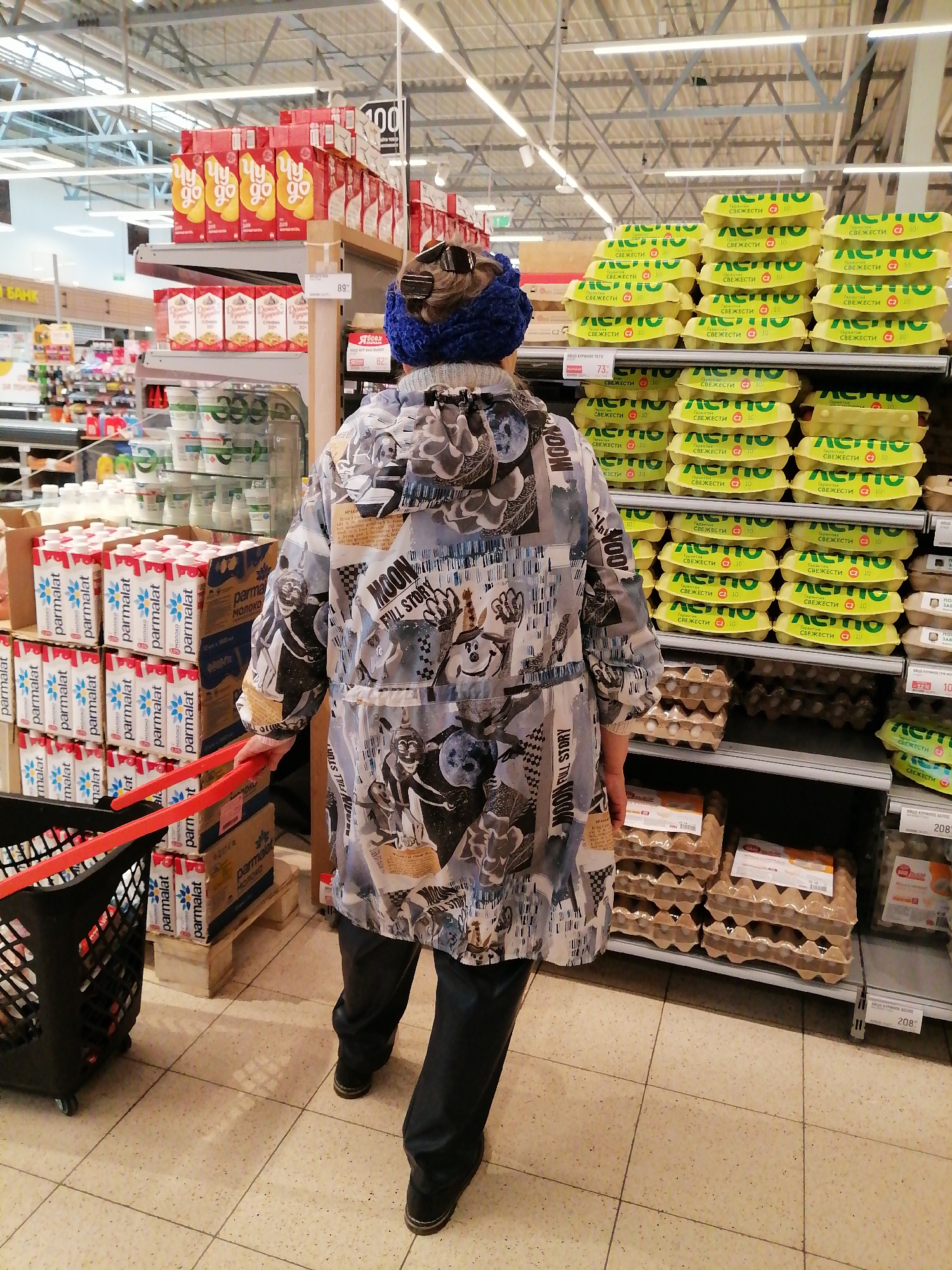 The image shows a person, viewed from the back, in a grocery store aisle. The person is wearing a blue knitted hat and a jacket with a collage-like pattern featuring cartoon characters, text, and images related to the moon. They are holding red handles of a shopping cart, which contains several cartons of milk. The shelves in the background are stocked with various products, including milk cartons, egg cartons, and other packaged goods. A price tag displaying "100" is visible on a shelf in the upper left. Another price tag shows "89" and "62" near some red boxes. The aisle is brightly lit, suggesting it is daytime. The overall scene depicts a typical shopping experience in a supermarket. Text visible includes brand names like "parmalat", "letov", and "Явсех", as well as phrases like "MOON FULL STORY" and "СВЕЖЕСТИ" (freshness).