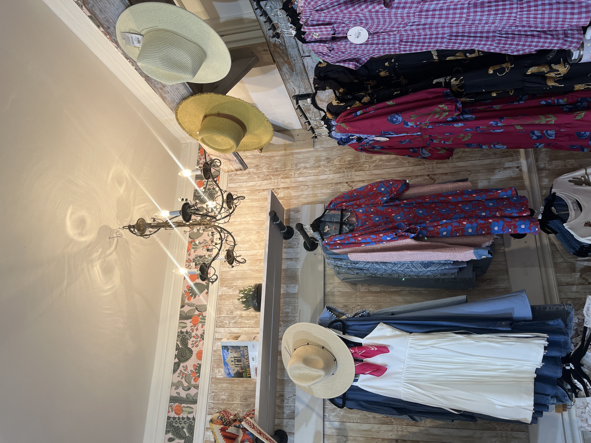 The image shows the interior of a retail store in New Braunfels, Texas. Clothing items and straw hats are displayed on racks and shelves. A chandelier is visible in the foreground. The walls are decorated with floral patterned wallpaper.