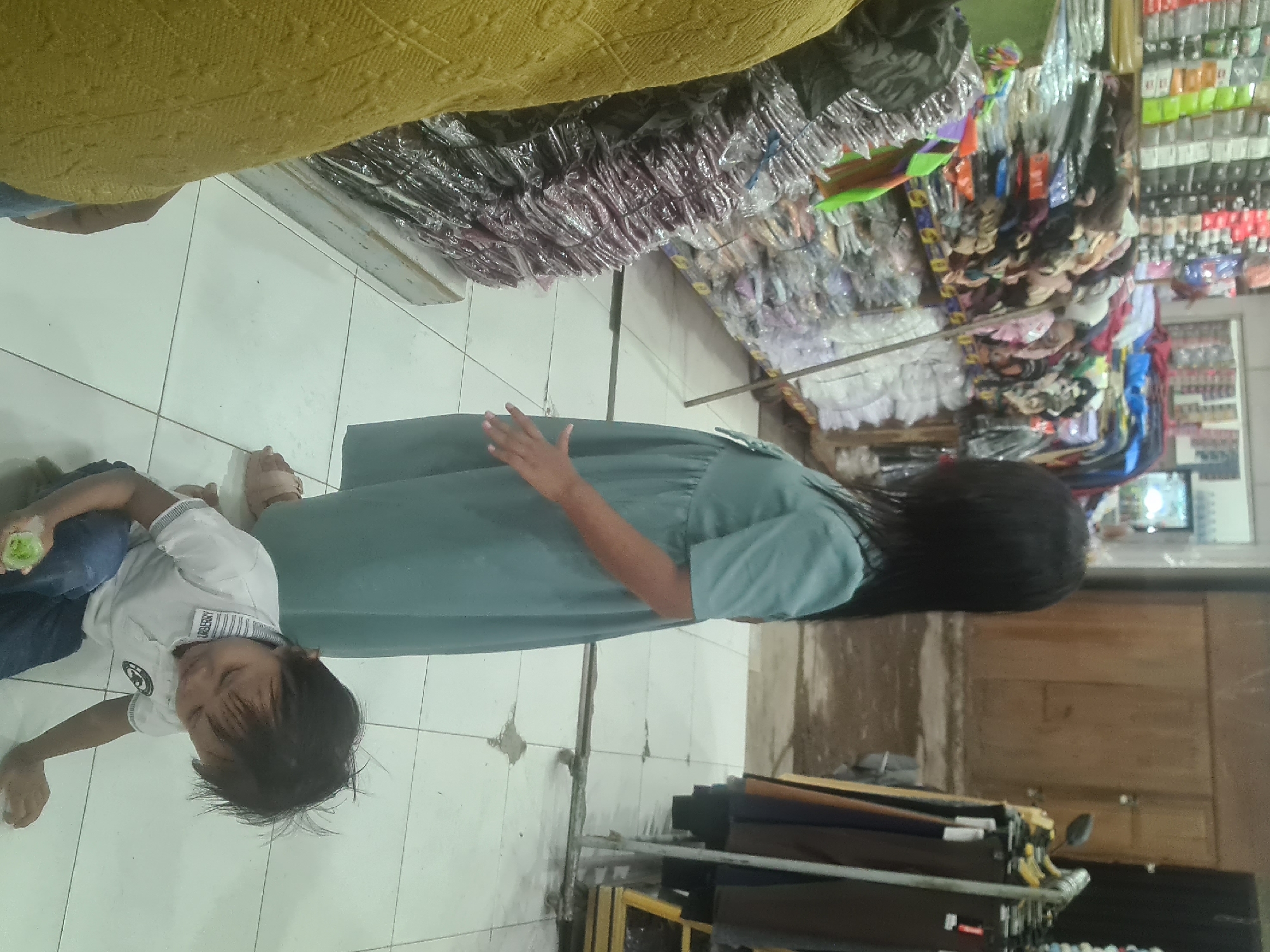 In a retail setting, possibly a clothing store in Lhokseumawe, Indonesia, a young girl stands near a display of packaged clothing. She has dark hair and wears a light blue dress. In the foreground, a young boy sits on the floor, wearing a white polo shirt and blue jeans. He holds a green object, possibly a piece of fruit or vegetable, in his left hand. The floor is tiled, and stacks of packaged garments are visible on shelves to the left. Clothing racks with garments are to the right. The overall environment appears to be an indoor market or shop.