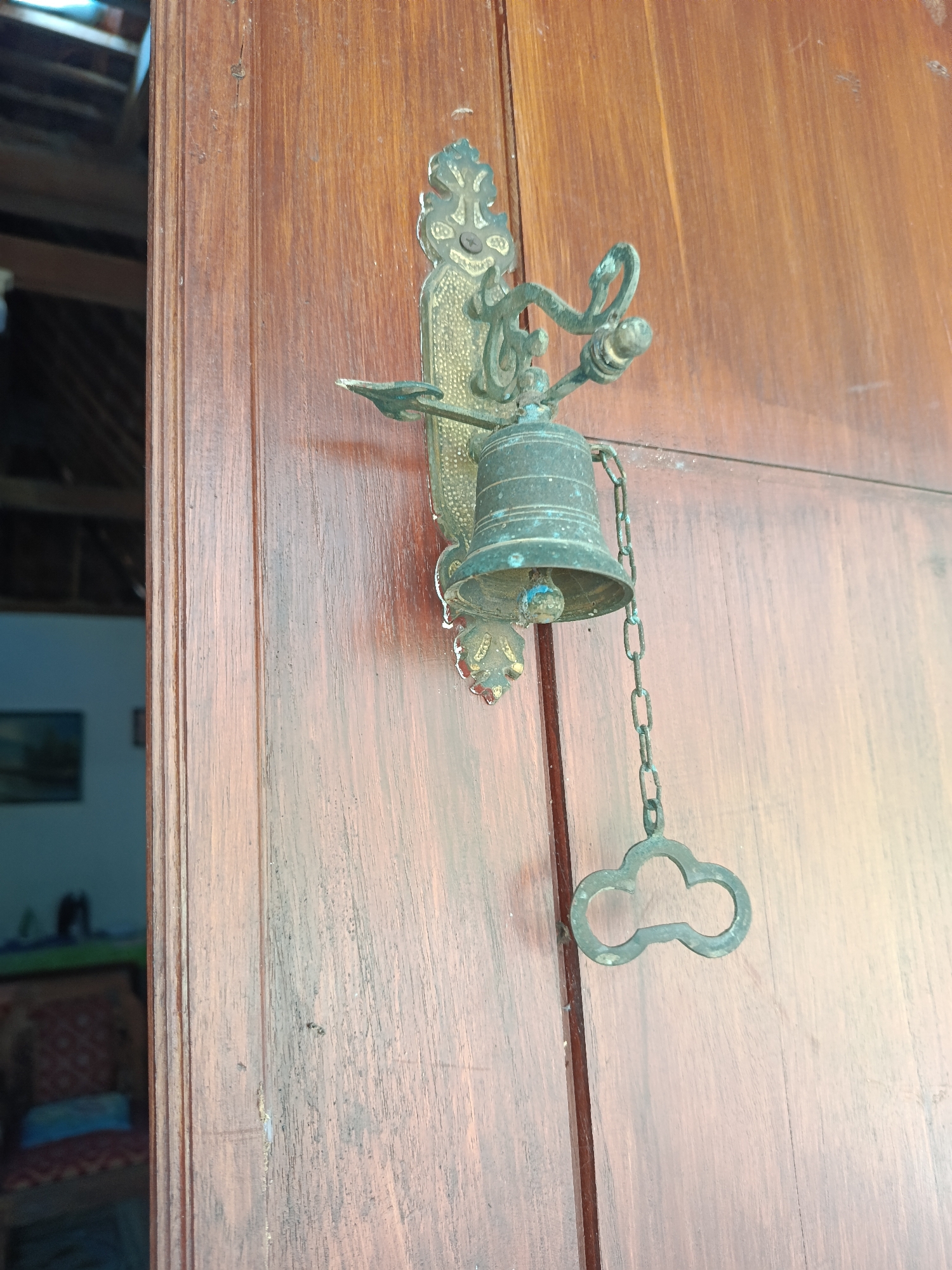 An antique brass door bell is mounted on a wooden door in a building. The bell is circular and suspended by a chain, with an ornamental handle attached to the chain. The door is made of dark wood with visible grain. In the background, a dimly lit interior can be seen, with furniture and a painting on the wall. The location is identified as Karangmojo, Indonesia. The image is taken from a slightly upward angle, focusing on the bell and the door.