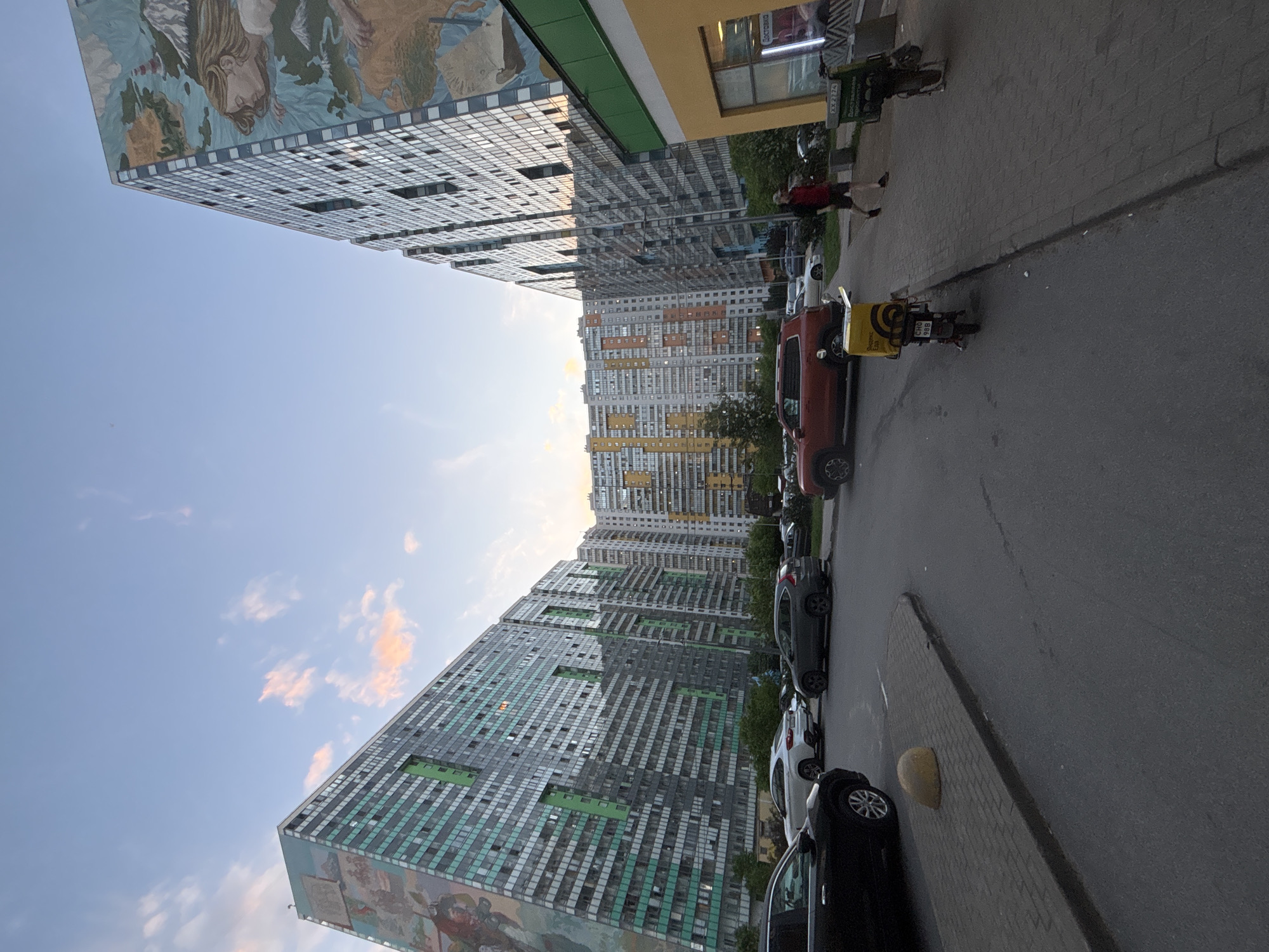 The image, taken with a vertical orientation (appears rotated 90 degrees counter-clockwise), captures a vibrant urban street scene dominated by modern, tall apartment buildings under a clear, late afternoon or early evening sky.
Two distinct high-rise buildings stand prominently on the left side. The closest building features a large, colorful mural on its side, depicting a stylized map or world with a woman's face and other figures. Its facade is composed of numerous square, reflective panels. Adjacent to it, another similar building displays green and grey/blue panels, with a mural also visible at its base. In the background, a cluster of other multi-story apartment blocks with varied colored facades (yellow, orange, grey) recede into the distance, creating a dense urban landscape.
On the right side of the image, a paved road runs parallel to the buildings, lined with parked cars. A red car is visible in the mid-ground, followed by a motorcycle or scooter equipped with a bright yellow delivery box. The text "Яндекс Еда" (Yandex Food) and "СПО 988" are clearly visible on the delivery box. Further down the street, several other cars, including dark grey, black, and white models, are parked.
A person wearing a red shirt is seen walking on the sidewalk, which features cobblestone paving, next to a yellow commercial building. Another person is partially visible near the motorcycle. A sign on the yellow building displays the word "ДОСТАВКА" (Delivery) and partial numbers "ХХХ 27-34". The sky overhead is blue with scattered white and light pink clouds, indicating either sunrise or sunset, with the warm light suggesting late afternoon or early evening. The overall impression is a bustling, modern residential area.
