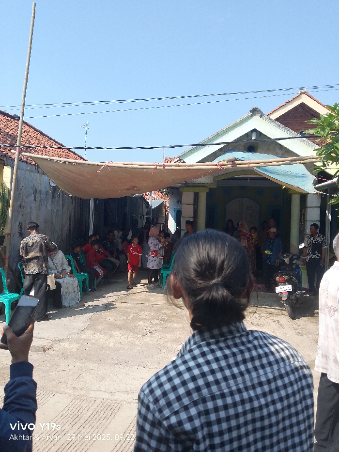 The image shows a gathering of people in an outdoor space, likely a courtyard or street, in Anjatan, Indonesia. There is a large awning stretched across the space, providing shade from the sun. Several people are sitting on chairs, while others are standing and interacting. A few children are playing in the space. There is a motorbike parked on the right side of the image, which is also in the shade of the awning. The environment is generally clean and well-maintained, with a paved surface and some greenery in the background.