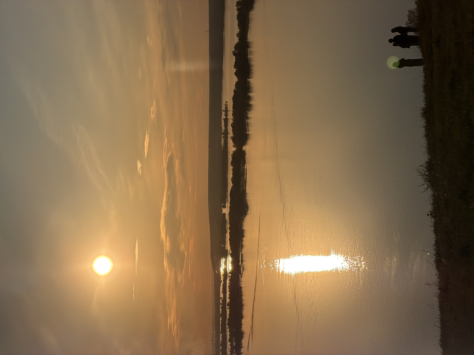 The image captures a sunset over a body of water, likely a river or lake, with the sun positioned on the left side of the frame. The sun's reflection creates a bright, elongated streak on the water's surface. Silhouettes of people are visible on a bank or embankment to the right. Trees or vegetation line the opposite shore in the distance. The sky is hazy with warm hues from the sunset, reflecting in the water. Given the location of Innopolis, Russia, the water body is most likely the Volga River or a reservoir connected to it. The people appear to be observing the sunset.