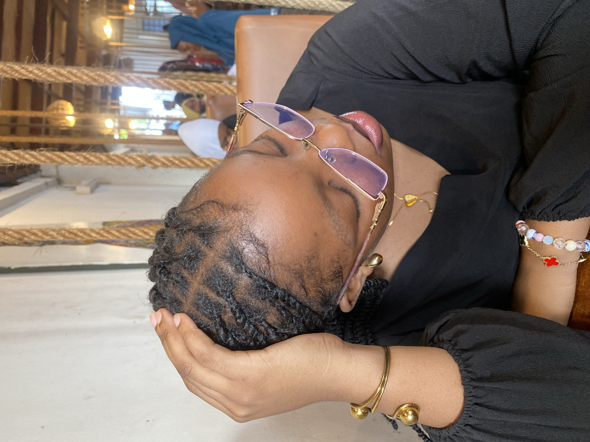 A woman rests her head on her hand while seated. She wears glasses with lavender-tinted lenses and gold frames. Her hair is braided, and she is wearing a black blouse, gold earrings, and multiple gold bracelets on her left wrist. A gold necklace with a small heart pendant is visible. In the background, a person wearing a white face mask and others can be seen. The scene appears to be indoors, possibly in Dar es-Salaam, Tanzania, based on the light and general environment.