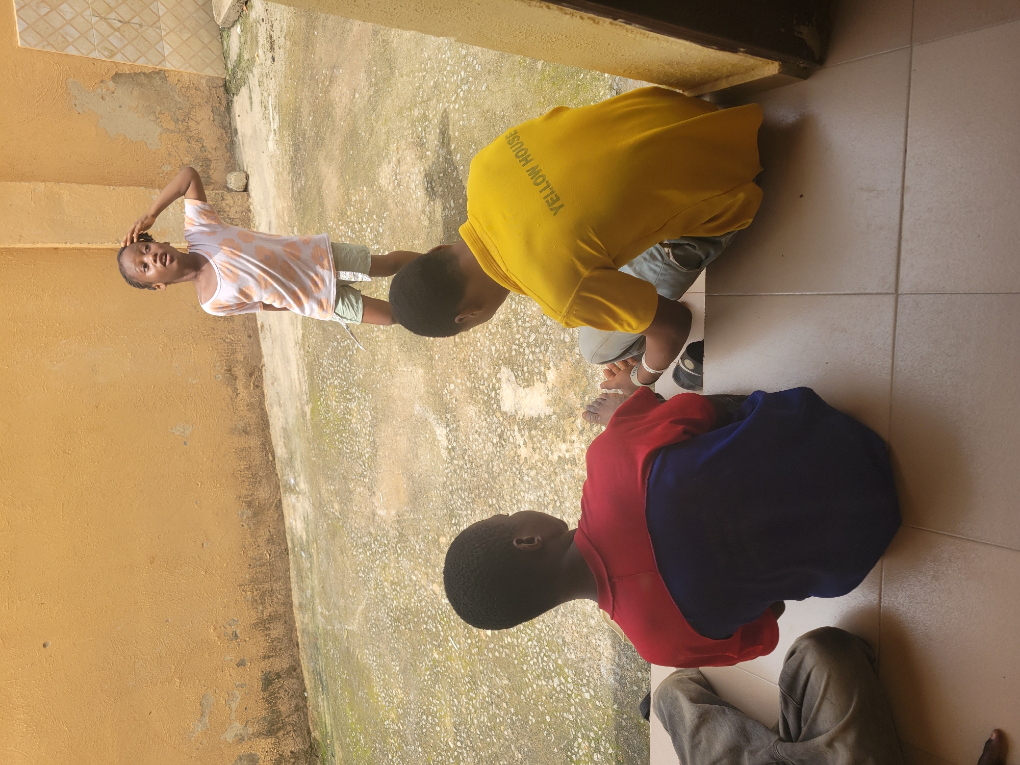 In an outdoor courtyard in Suleja, Nigeria, three young individuals are present. One child stands looking upwards with their hand touching their head, wearing a light-colored t-shirt with an orange floral pattern and light-colored shorts. Two other children are seated on the ground. One is facing away from the camera, wearing a t-shirt with red and blue sections. The other child, also facing away, is wearing a bright yellow t-shirt with the words "YELLOW HOUSE" printed on the back in dark text. The ground is paved with a rough, speckled concrete material, and a textured, orange-painted wall forms the background. The scene appears to be in front of a doorway or entrance.