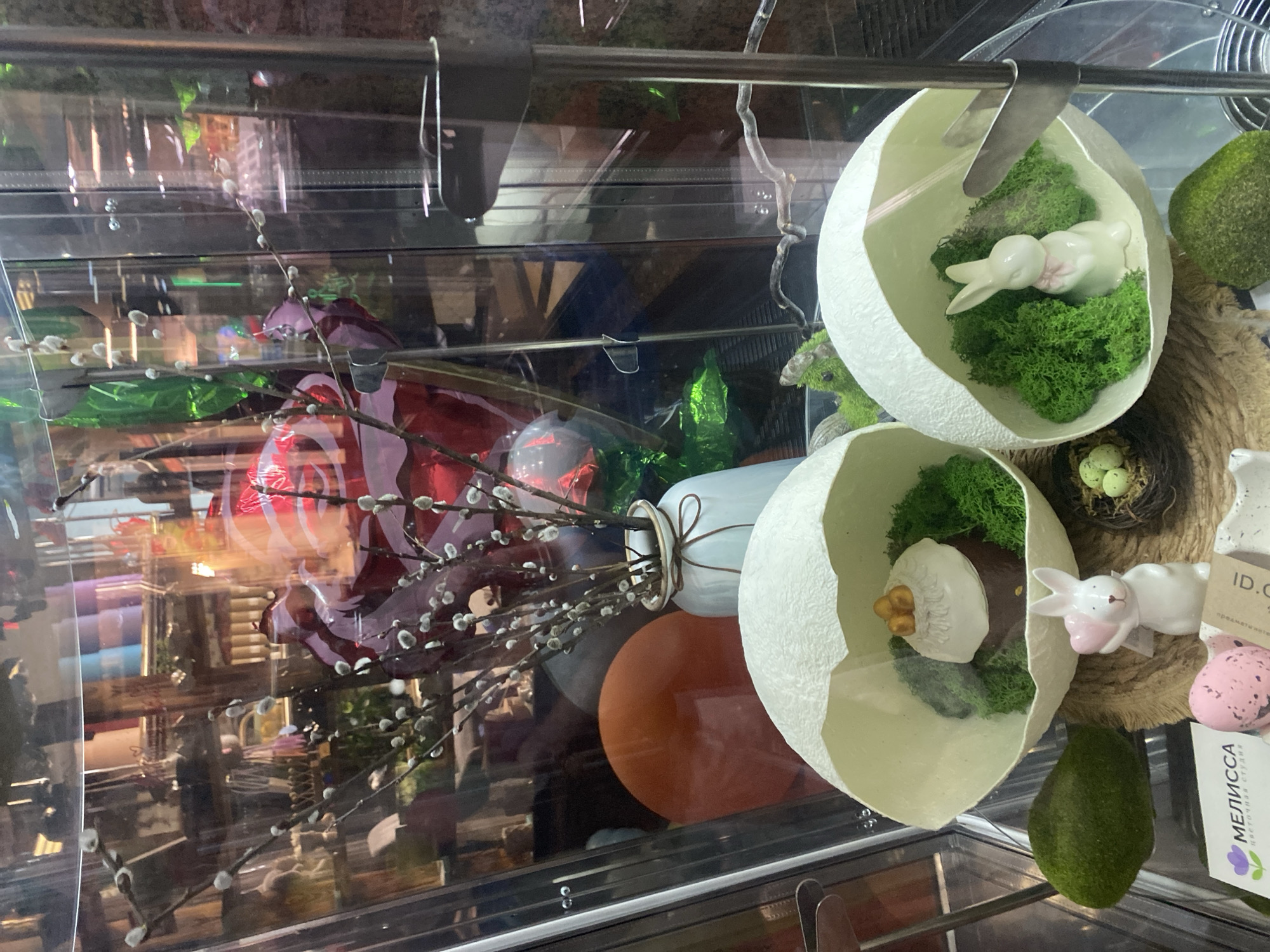 The image shows a close-up view of Easter decorations in a glass display case. The decorations include two halves of a white eggshell, each containing artificial green moss and a small, white ceramic bunny. There is a small nest with green eggs. Other decorative elements visible are pussy willow branches in a white vase, colorful wrapped candies, and additional mossy rocks. A business card for "MELISSA" is visible. The location is implied to be Sochi, Russia based on the text on the card.