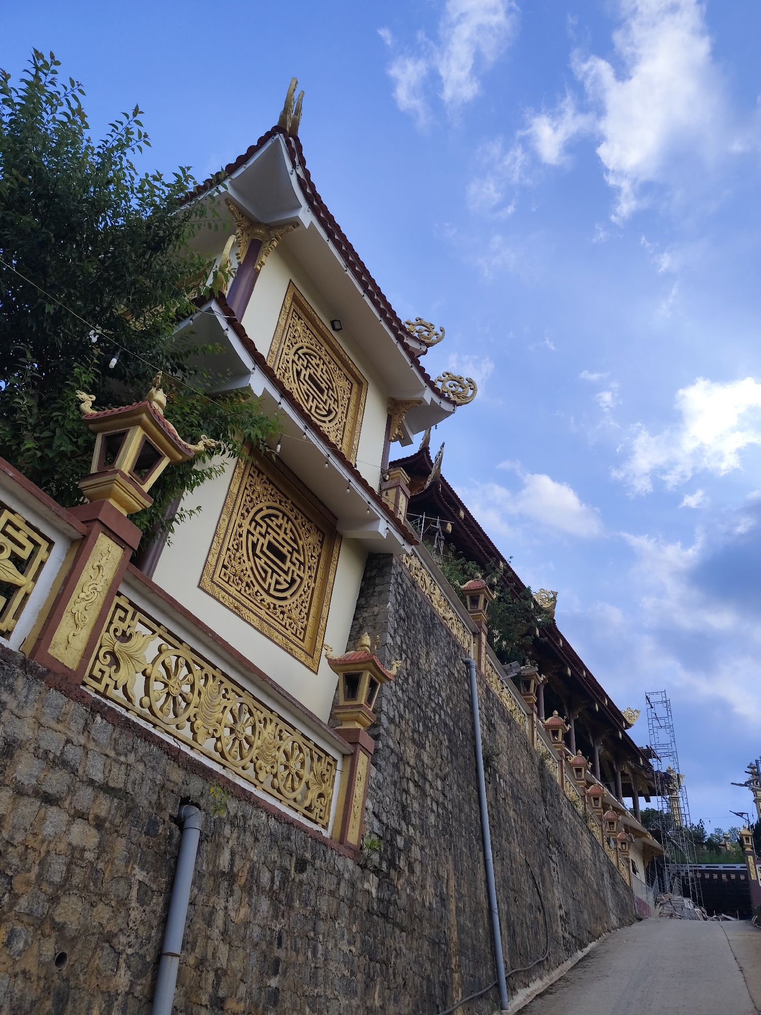 The image shows a building in Lạc Dương, Vietnam, specifically a Buddhist temple. The architectural style is Vietnamese, featuring a multi-tiered structure, a red-tiled roof, and ornate gold decorations. The building is built on a stone wall that is alongside a road. A gray PVC pipe runs down the wall. There is scaffolding on the building, indicating construction or renovation. A tree is visible, and the sky is blue with scattered clouds.
