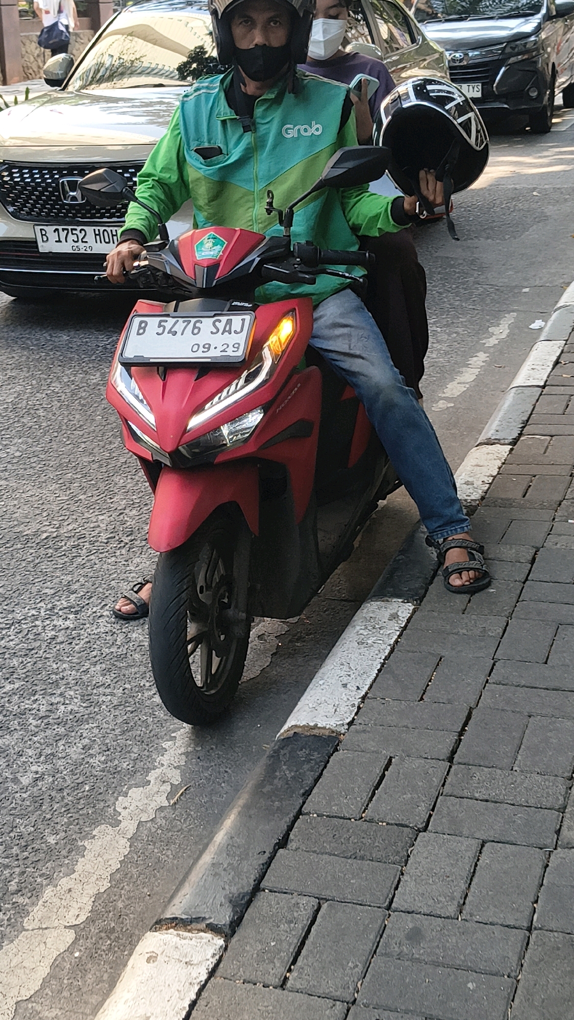 An outdoor, eye-level medium shot captures a man wearing a green and teal Grab driver's uniform, a black helmet, and a black face mask, sitting on a red scooter in South Jakarta, Indonesia. The scooter's license plate displays "B 5476 SAJ" and "09.29". The driver holds a black helmet in his right hand. Behind him, a woman wearing a white mask and a purple shirt is visible. A white car with the license plate "B 1752 HOH" and "05.29" is parked behind the scooter. The scene is set on a street with a paved sidewalk to the right, bordered by a curb. The road surface is asphalt with visible white paint markings. The lighting suggests it is daytime.
