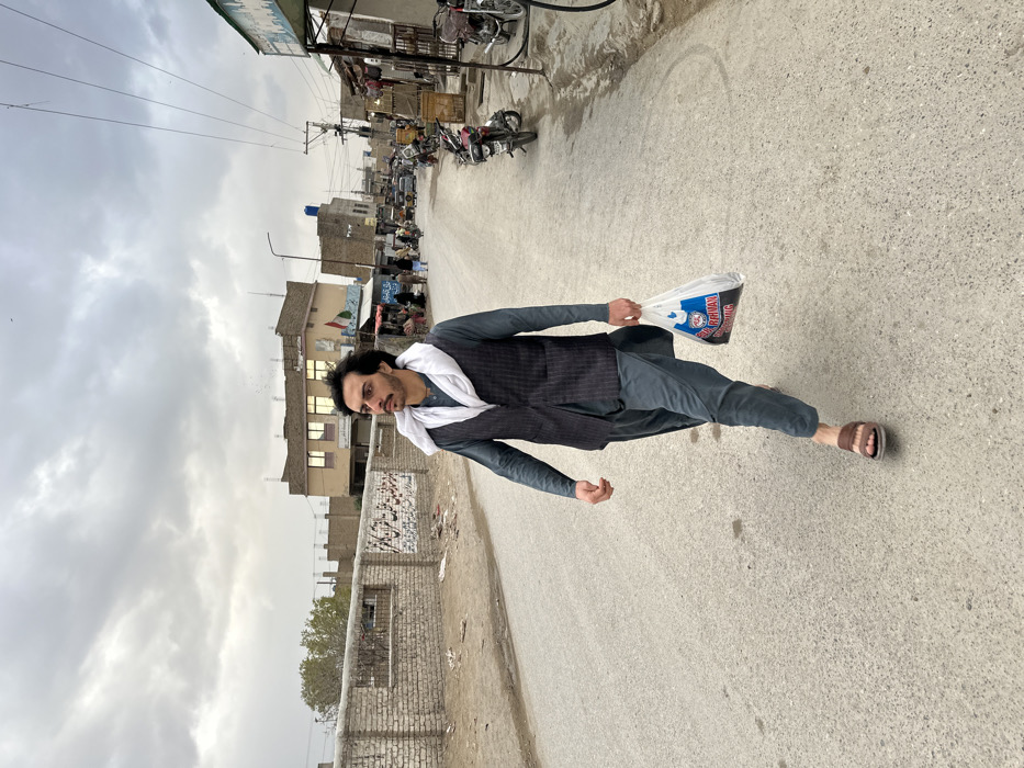 The image portrays an urban street scene within Quetta City Tehsil, Pakistan, under an overcast sky. In the foreground, a male subject, approximately 20-30 years old, with dark hair, a mustache, and a short beard, walks across a grey asphalt road. He wears a dark grey long-sleeved tunic (likely a shalwar kameez), a dark pinstriped vest, and brown sandals. A white cloth is draped around his neck and shoulders. He holds a white plastic shopping bag with blue and red text in his left hand, looking slightly upwards and to his right.
The left side of the street features a low brick wall with visible script, followed by an area with a sparse tree and several tan-colored buildings. The right side of the street is a busy thoroughfare with multiple indistinct individuals, numerous parked motorcycles, additional vehicles further down the road, utility poles, and overhead power lines. A row of commercial buildings with awnings and signs lines this side. The environment is consistent with a populated urban area, with some debris visible along the roadside.