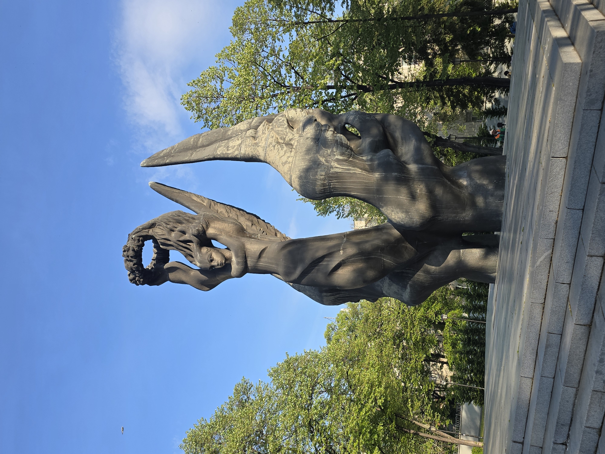 The image shows a bronze statue in a park on a sunny day. The statue is of a woman with wings, holding a wreath above her head. The trees are green and lush, and the sky is blue with some white clouds. The statue is located in Unknown, Unknown.