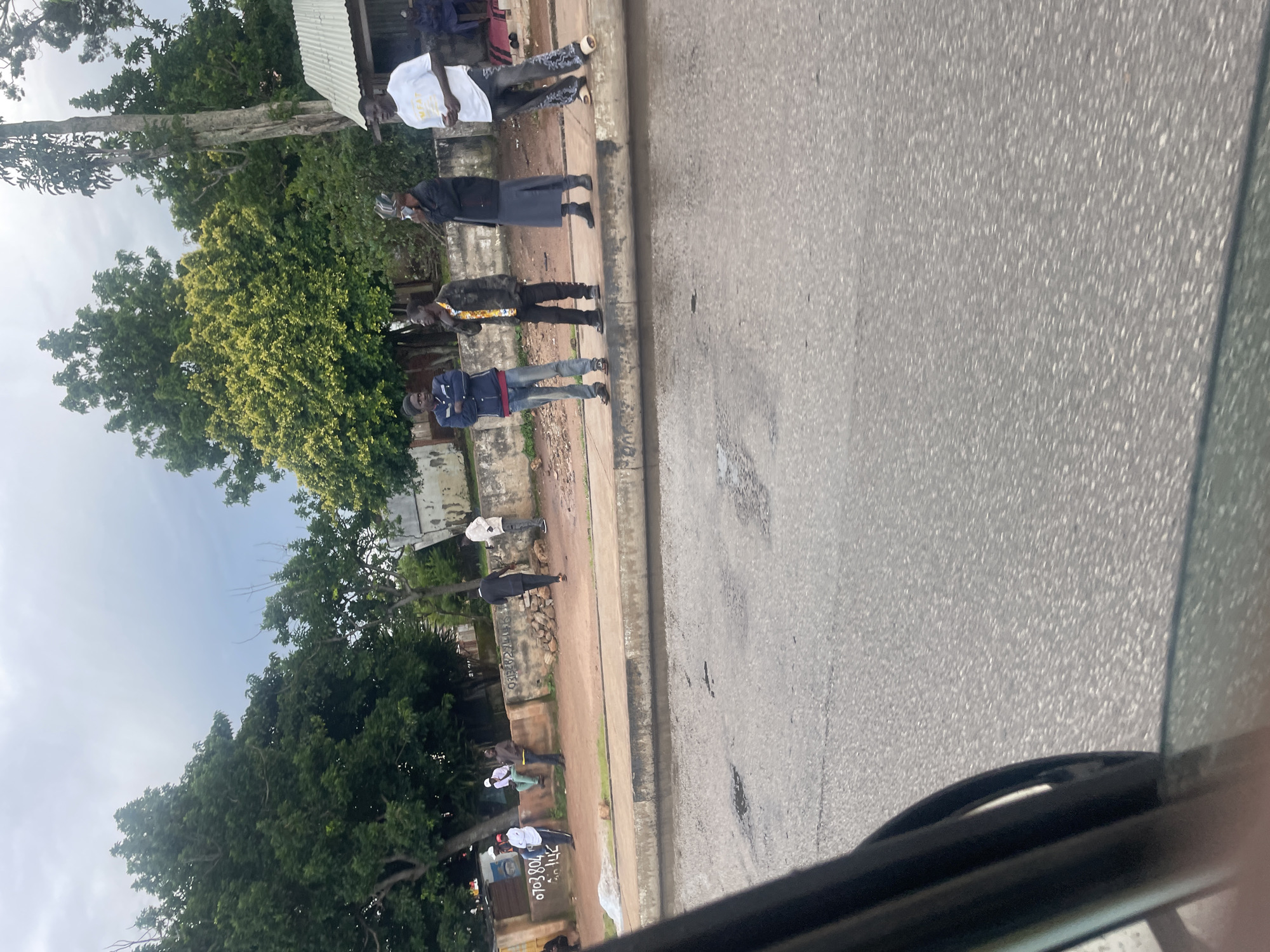 This is a medium shot taken from inside a vehicle, looking out at a street scene in Jos, Nigeria. A group of men are standing on a sidewalk next to a road. In the foreground, the edge of a car's window and side mirror is visible on the left.
On the sidewalk, from left to right, there are five men. The first man on the left is wearing a blue jacket, a red shirt, and blue jeans. The second man is wearing a camouflage jacket and black pants. The third man is wearing a black jacket and a long grey skirt or robe, and he appears to be on his phone. The fourth man is wearing a white t-shirt with "MEAT" printed on it and patterned pants. The fifth man is further to the right and is partially obscured, wearing a white garment. Behind the men, there is a low wall with some graffiti, followed by trees and buildings. The sky is overcast. The road in front of the sidewalk is paved and appears to have some puddles.