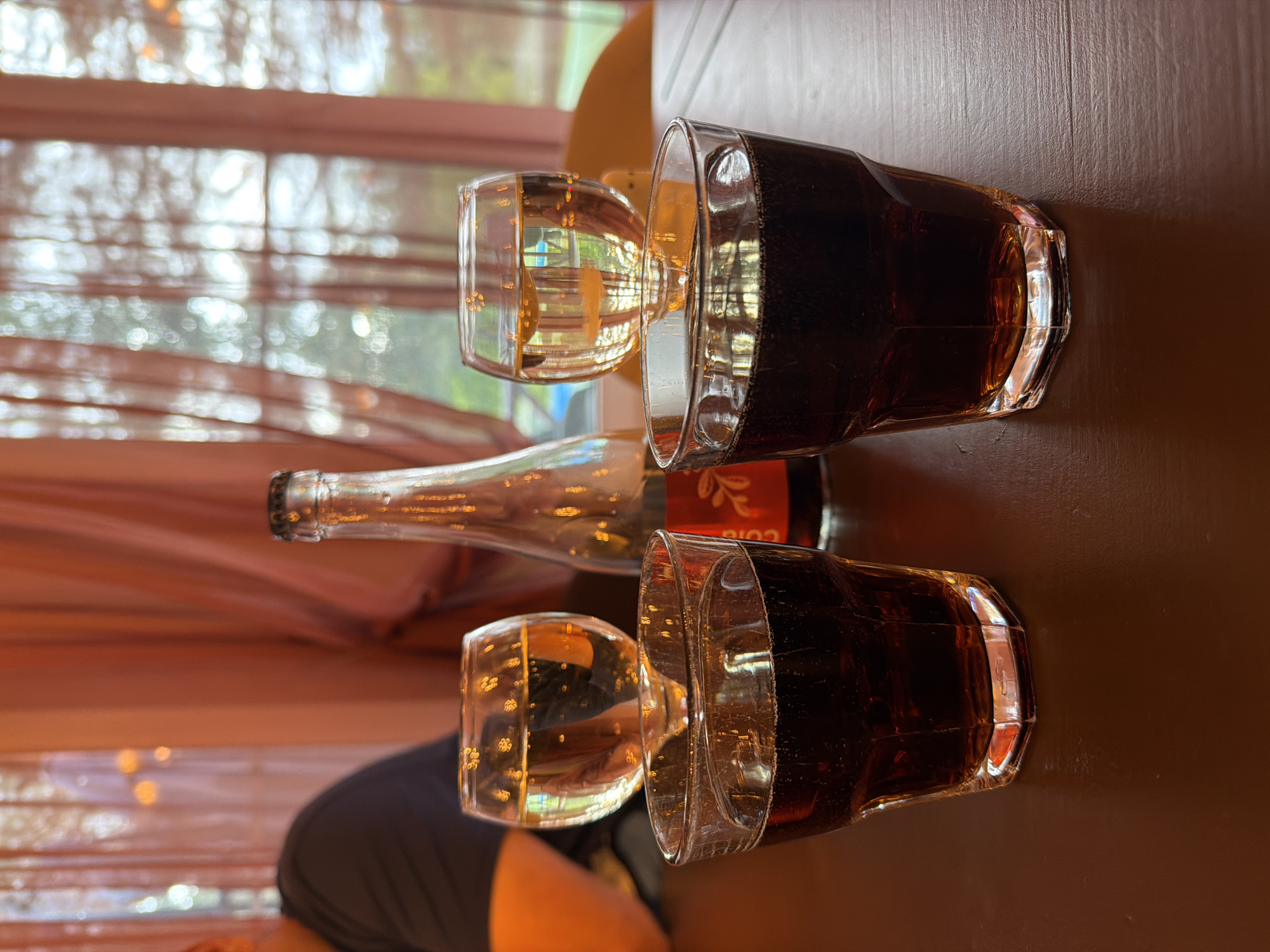 On a brown tabletop are two pairs of glasses and a glass bottle. One glass in each pair contains a dark liquid, while the other contains a clear liquid. The bottle appears to be empty. The location is indoors, possibly near a window with blinds or sheer curtains, with glimpses of the outside. Zaplav'e, Russia, is not verifiable from the image.